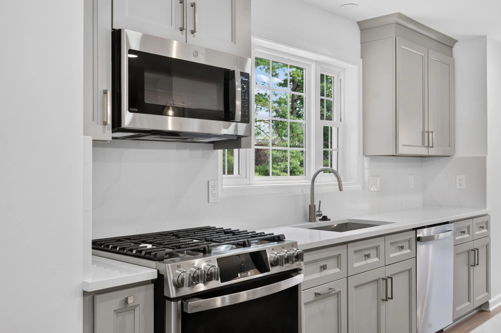 Modern kitchen with white cabinets, stainless steel microwave, gas stove, and large window showing green trees outside.