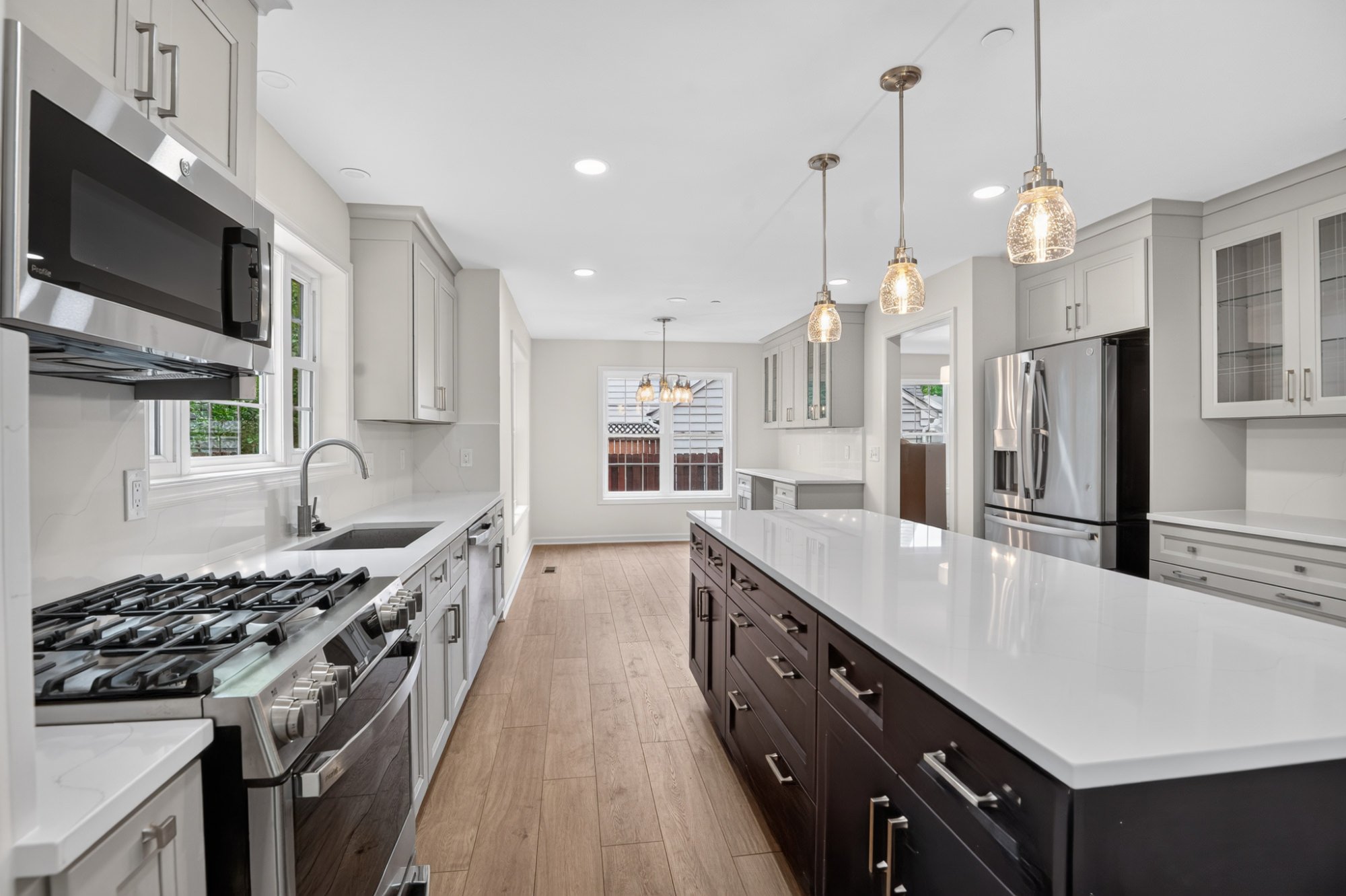 Modern kitchen with white and dark cabinets, stainless steel appliances, pendant lighting, hardwood floors, and windows showing outdoor greenery.