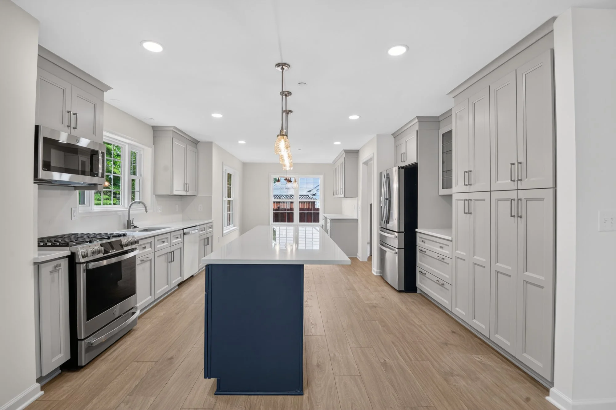 Modern kitchen with light gray cabinets, stainless steel appliances, a white countertop island with blue base, wood flooring, and large windows providing natural light.