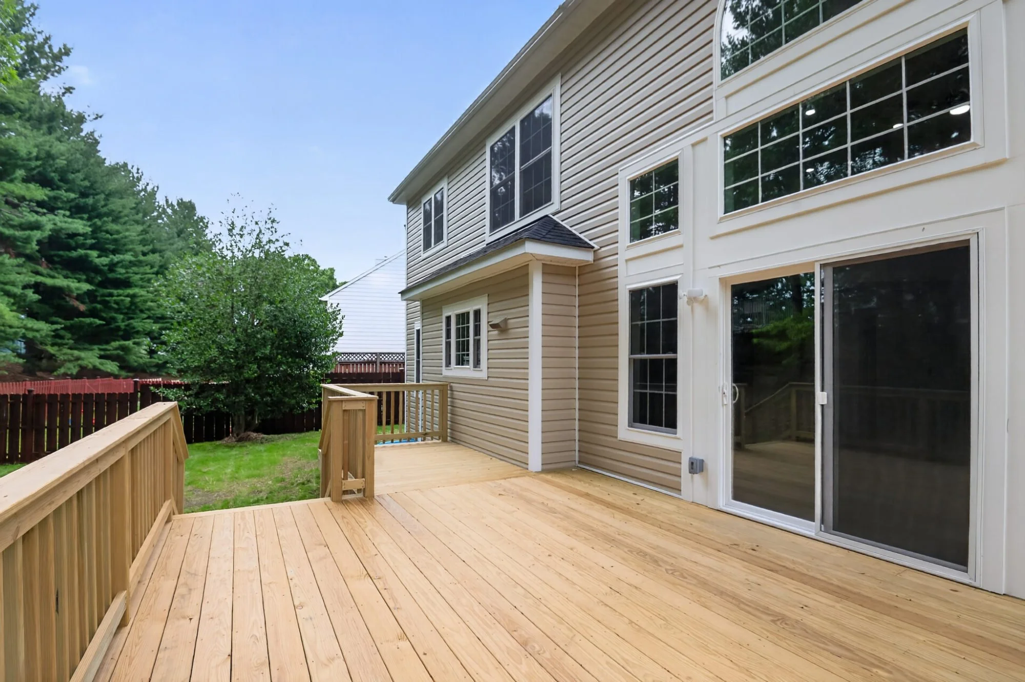 Backyard view with a wooden deck attached to house, sliding glass door, green grass, trees, and a fence in the background.