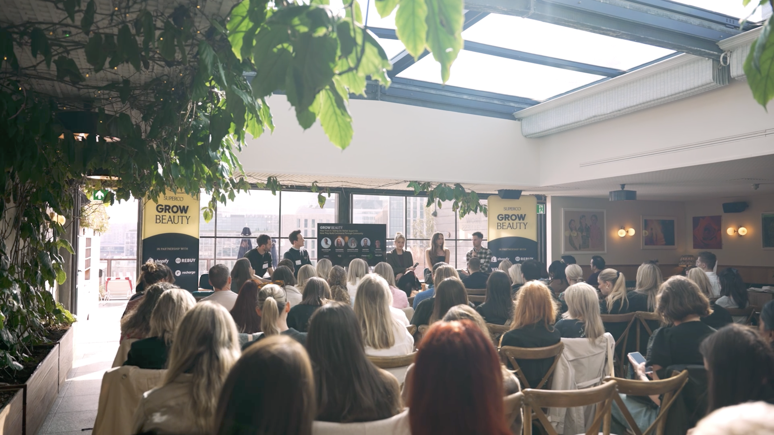 A large group of people attending a conference or seminar in a well-lit room with natural sunlight streaming through large windows and a glass ceiling. There are speakers or panelists at the front, and a presentation screen with the title 'Grow Beaut