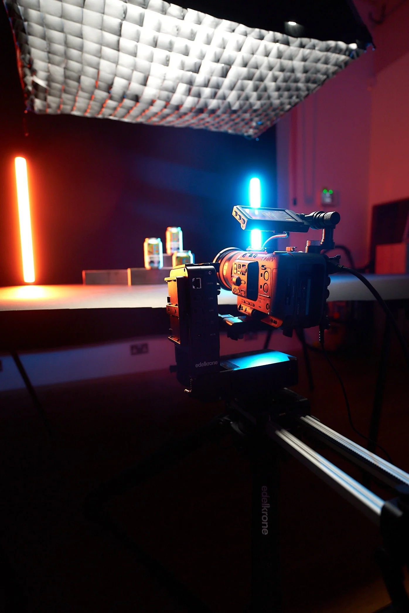 A professional video camera mounted on a rig in a studio setup, with colorful LED lights illuminating a table with cans, and a geometric ceiling pattern overhead.