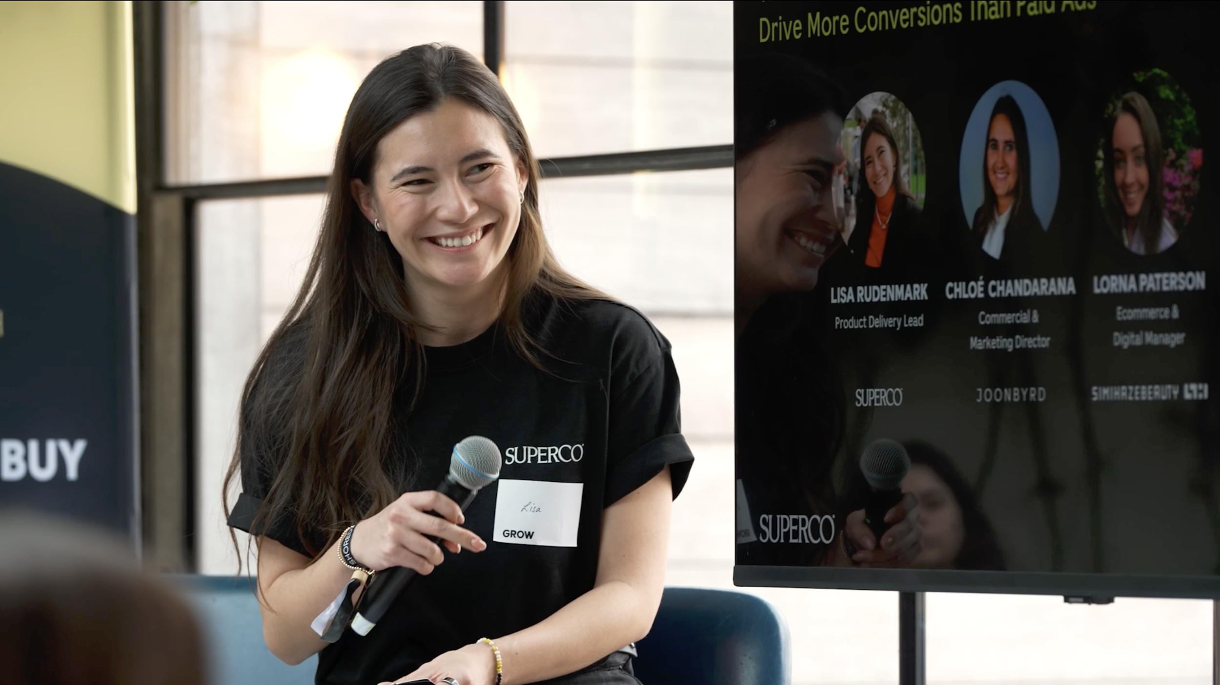 A woman with long brown hair, wearing a black t-shirt with a name tag labeled 'Lisa' and 'GROW,' is smiling and holding a microphone. She is sitting in front of a presentation screen with her reflection visible on it. The screen displays headshots an
