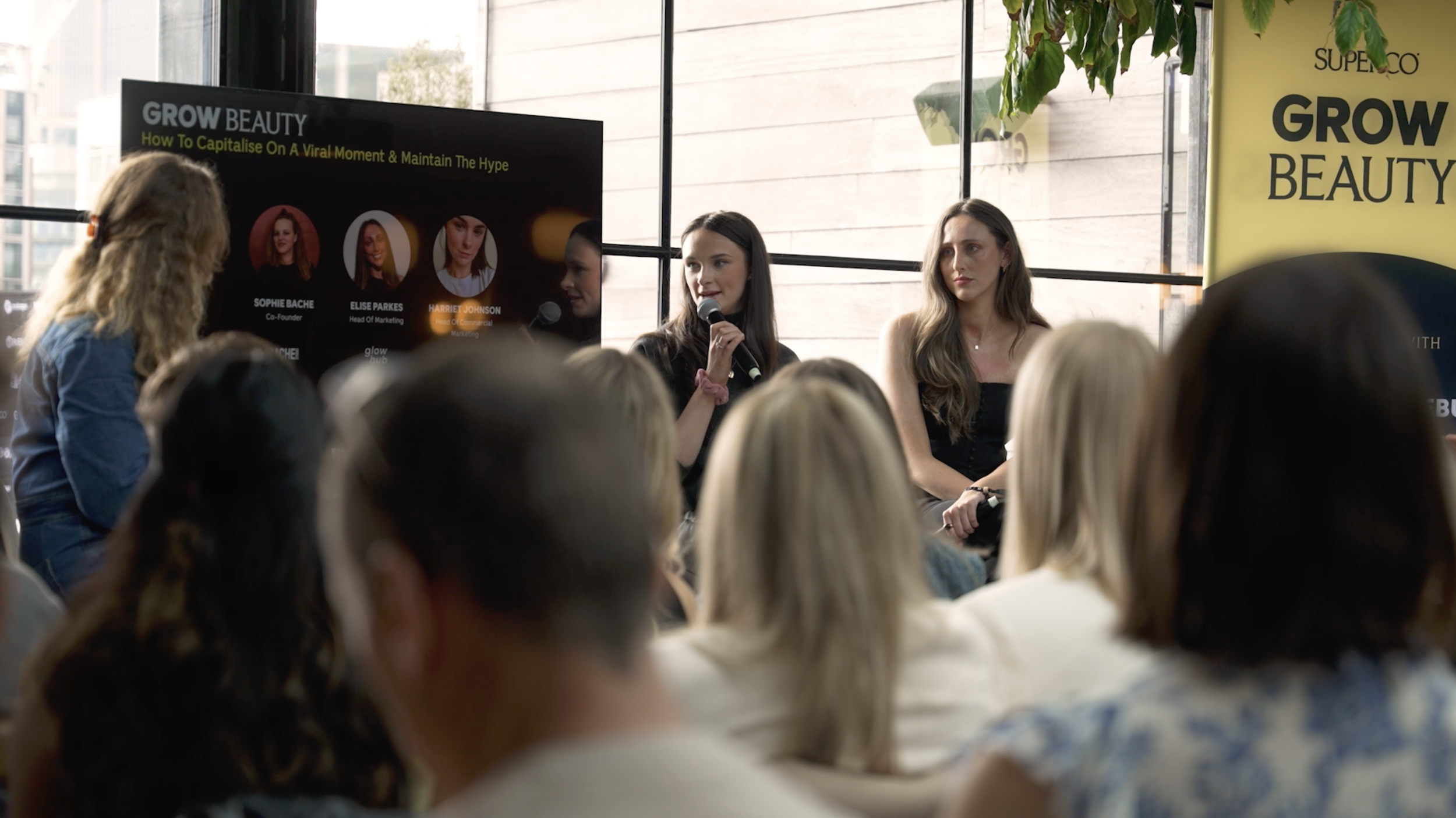 A woman with dark hair holding a microphone speaking at a panel discussion at an event titled GROW BEAUTY, with a screen displaying her name and title, and an audience listening attentively.