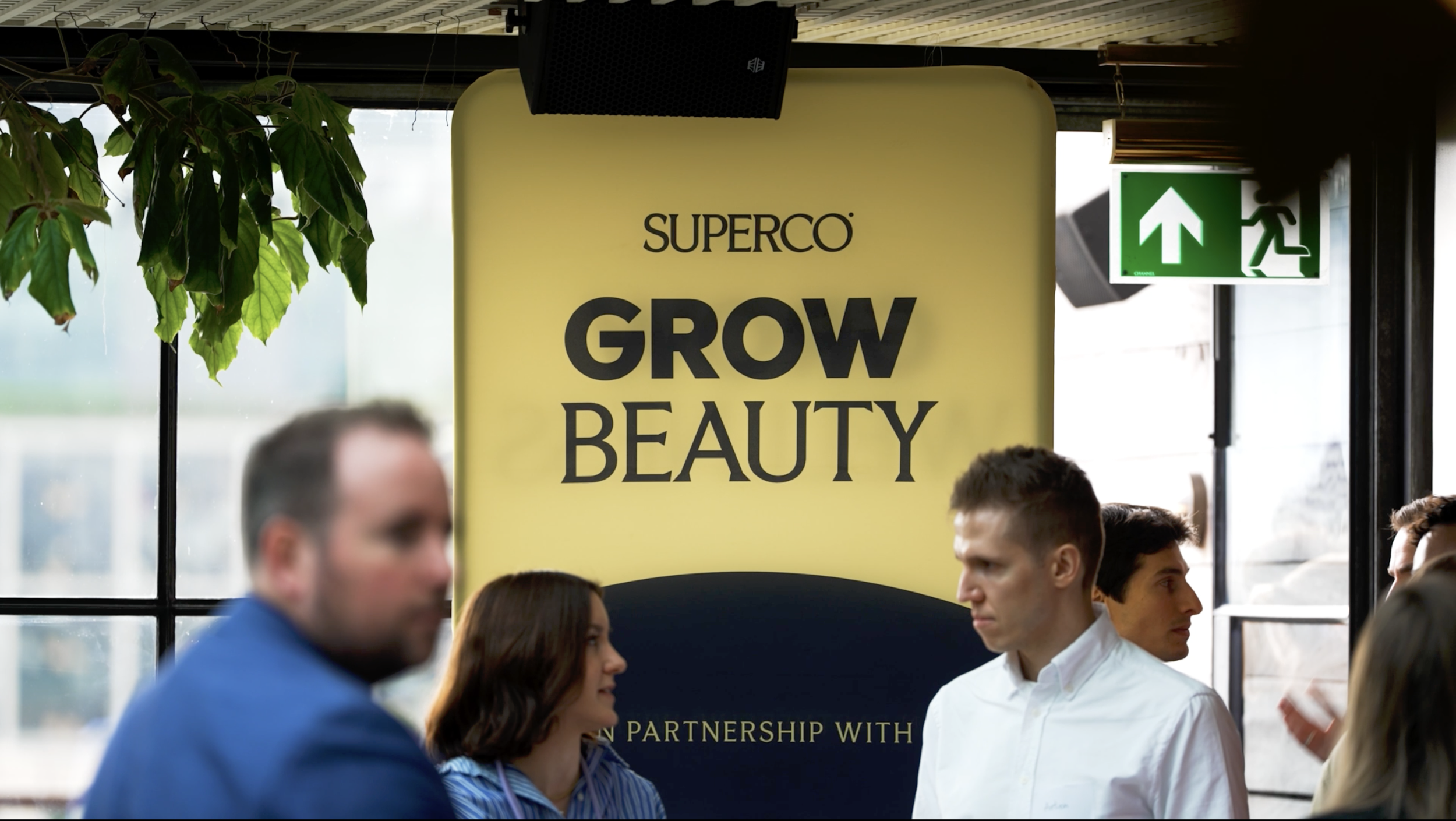 A promotional sign for SuperCo's Grow Beauty event or campaign. The sign features the text 'GROW BEAUTY' prominently displayed, with other smaller text not fully visible, inside a location with several people and an exit sign visible in the backgroun