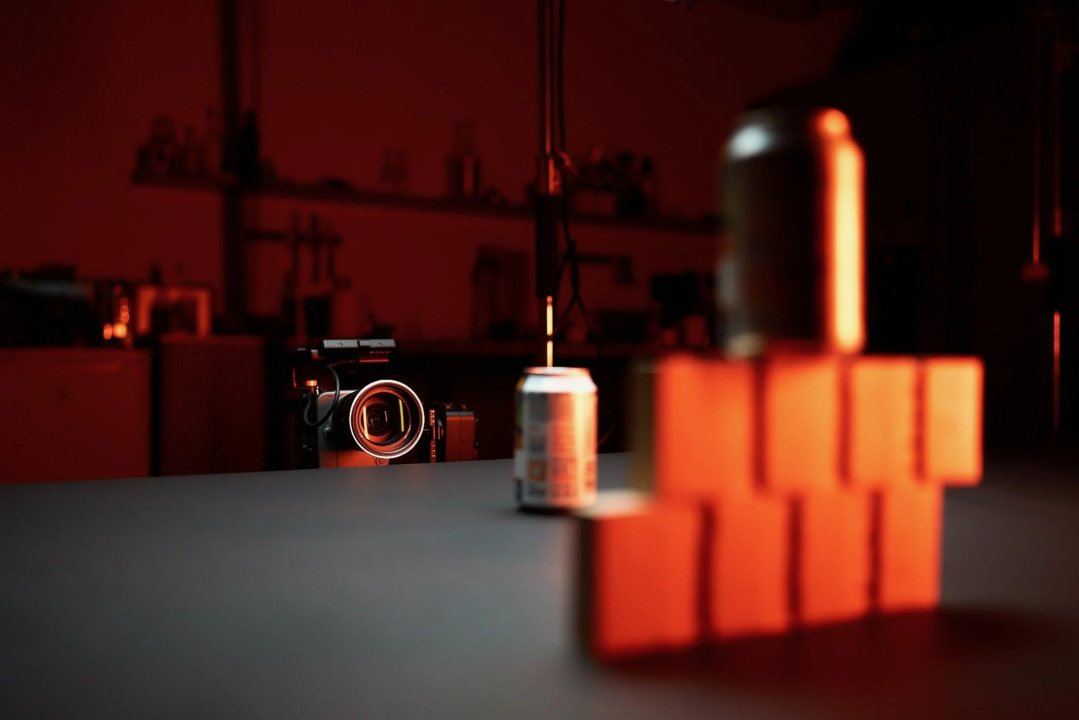 A camera on a table with a soda can, orange building blocks, and a bottle against a red background with shelves and equipment.