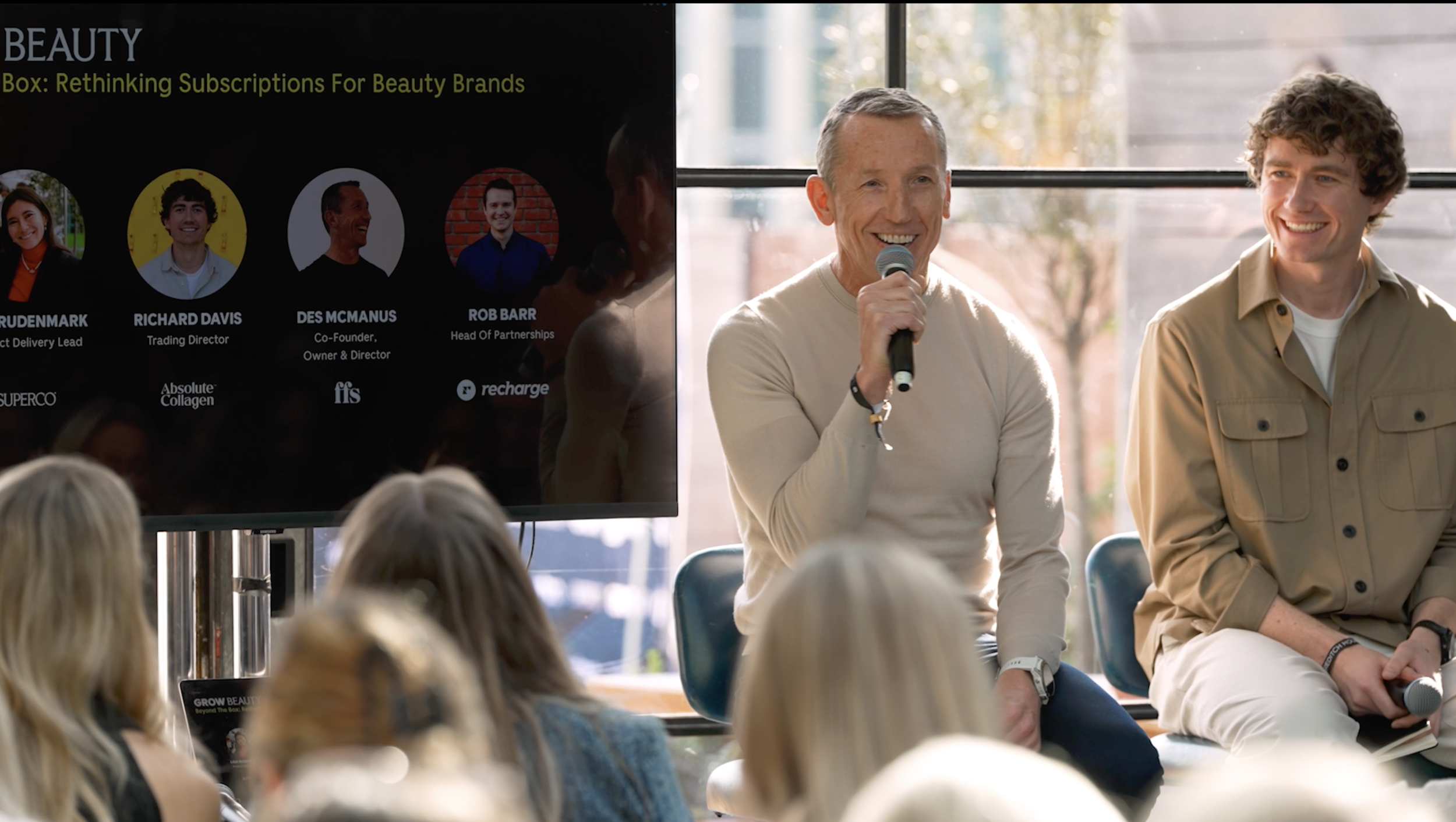 Two men seated on chairs in front of an audience, one speaking into a microphone, during a presentation on beauty brands with a large screen displaying profiles of speakers.