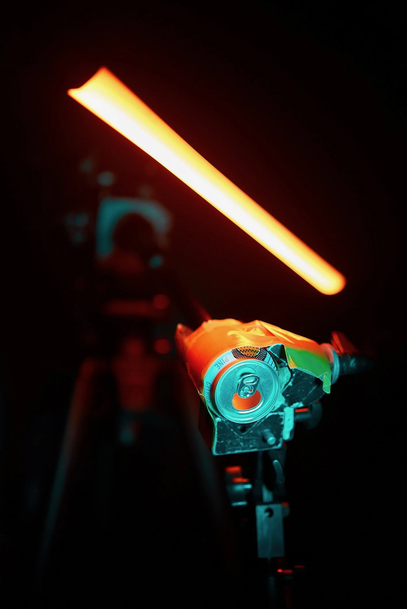 Close-up of a black gun barrel with a soda can wrapped in tape resting on top, illuminated by an orange laser beam in a dark background.