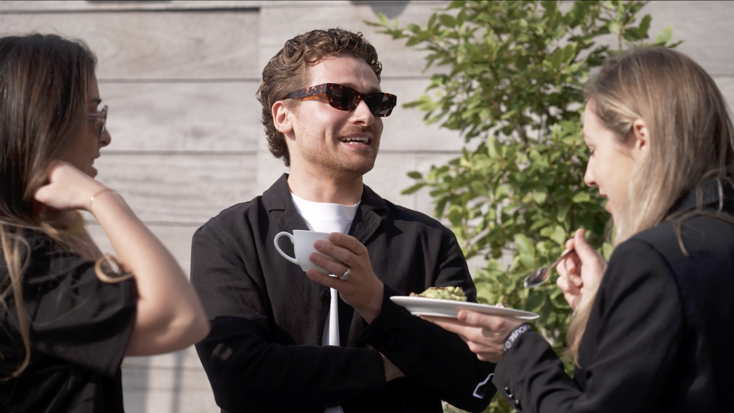 Three young adults having a conversation outdoors; one man holding a cup, one woman eating a spoonful of food, and another woman with glasses touching her hair, near a green bush and a wooden fence.