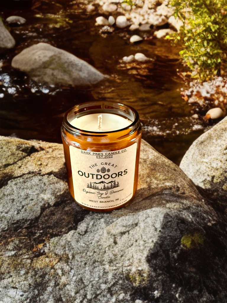 A lit candle in a brown glass jar with a label reading "The Great Outdoors," placed on a large rock near a flowing creek surrounded by rocks and greenery.