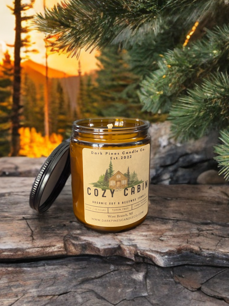 A brown jar candle labeled 'Cozy Cabin' by Dark Pines Candle Co. sits on a rustic wooden surface outdoors, with a pine branch in the background and a sunset sky.