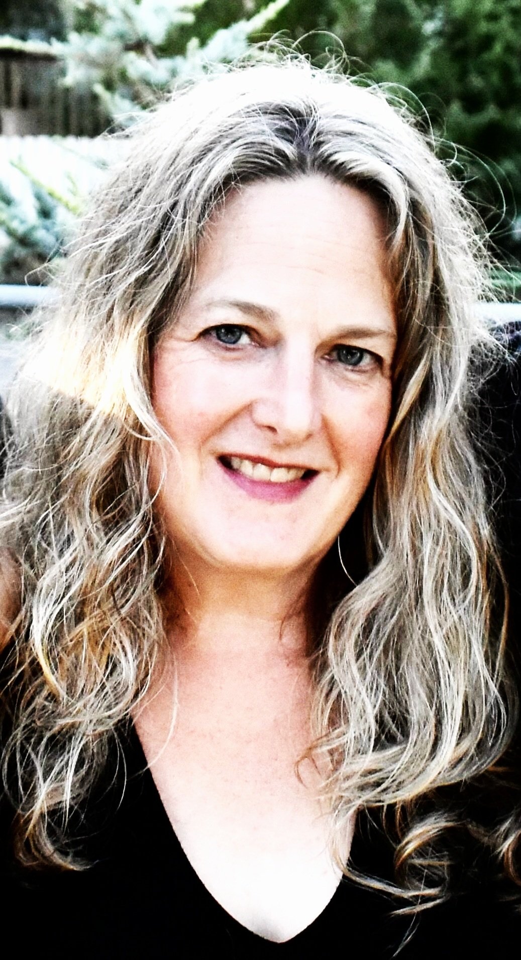 A woman with long, curly, blonde hair smiling outdoors with greenery in the background.