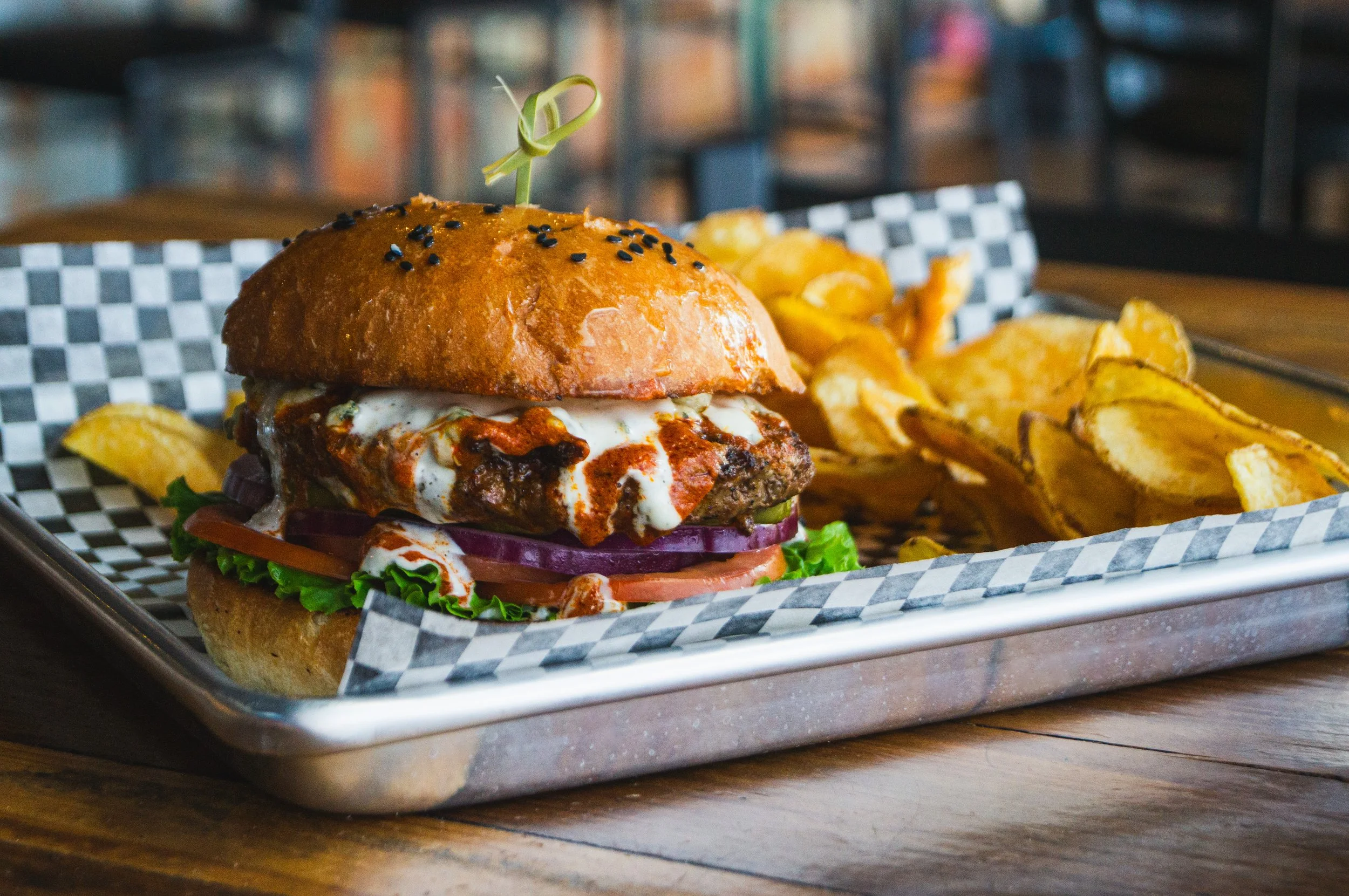 A gourmet burger with lettuce, tomato, onions, cheese, and sauce in a bun topped with black sesame seeds, served with a side of potato chips on a tray.