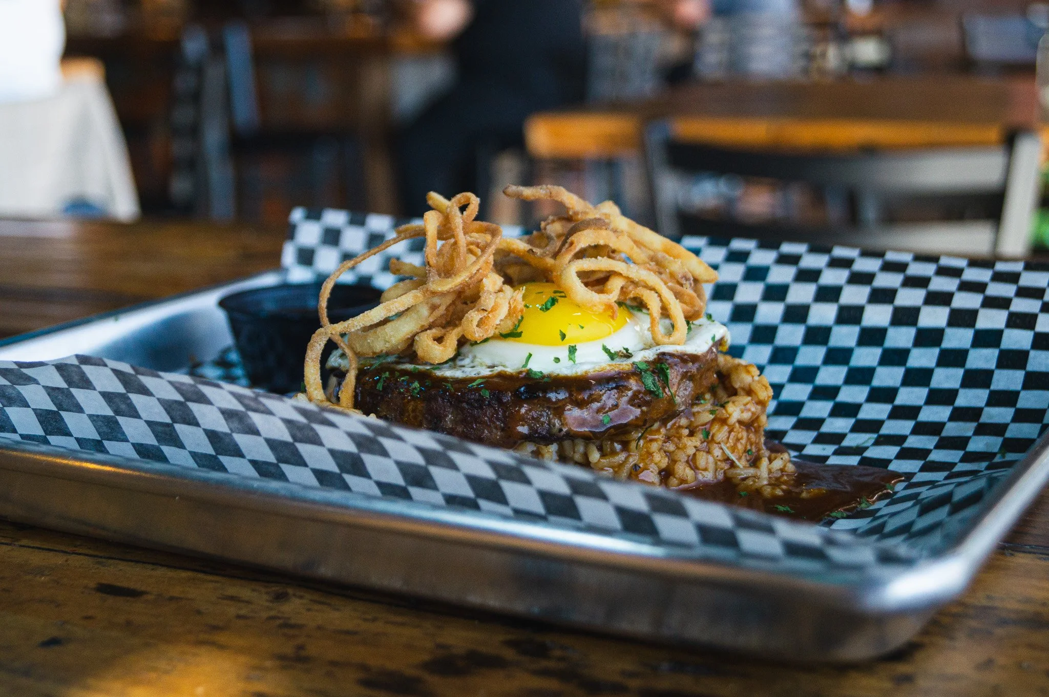 A serving of food on a checkered paper-lined tray featuring a fried egg on top, crispy fried onions, a beef patty, and a layer of rice with a side of dark sauce in a black cup, on a wooden table in a restaurant.