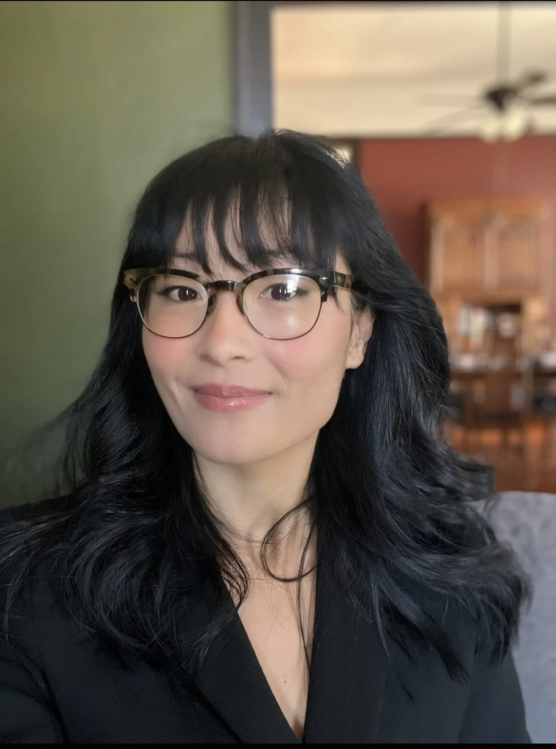 A woman with shoulder-length black hair, wearing glasses, smiling, in an indoor setting with wooden furniture in the background.