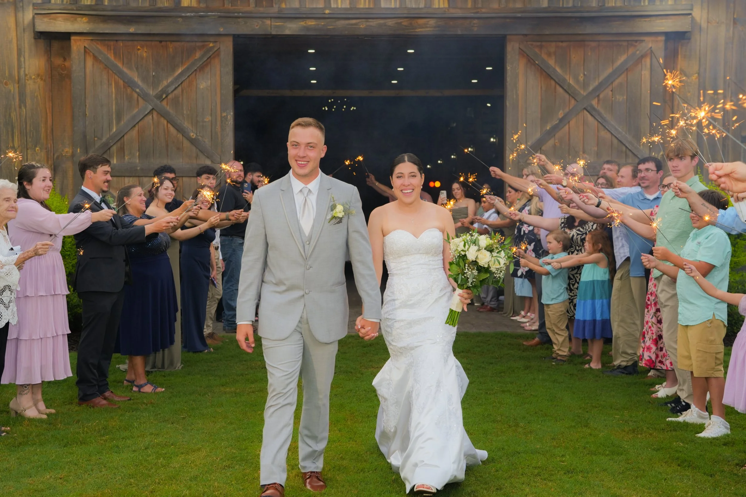 A newlywed couple walking hand-in-hand through a line of guests holding sparklers during an outdoor wedding celebration at night. The bride is in a white wedding dress holding a bouquet, and the groom is in a light gray suit.