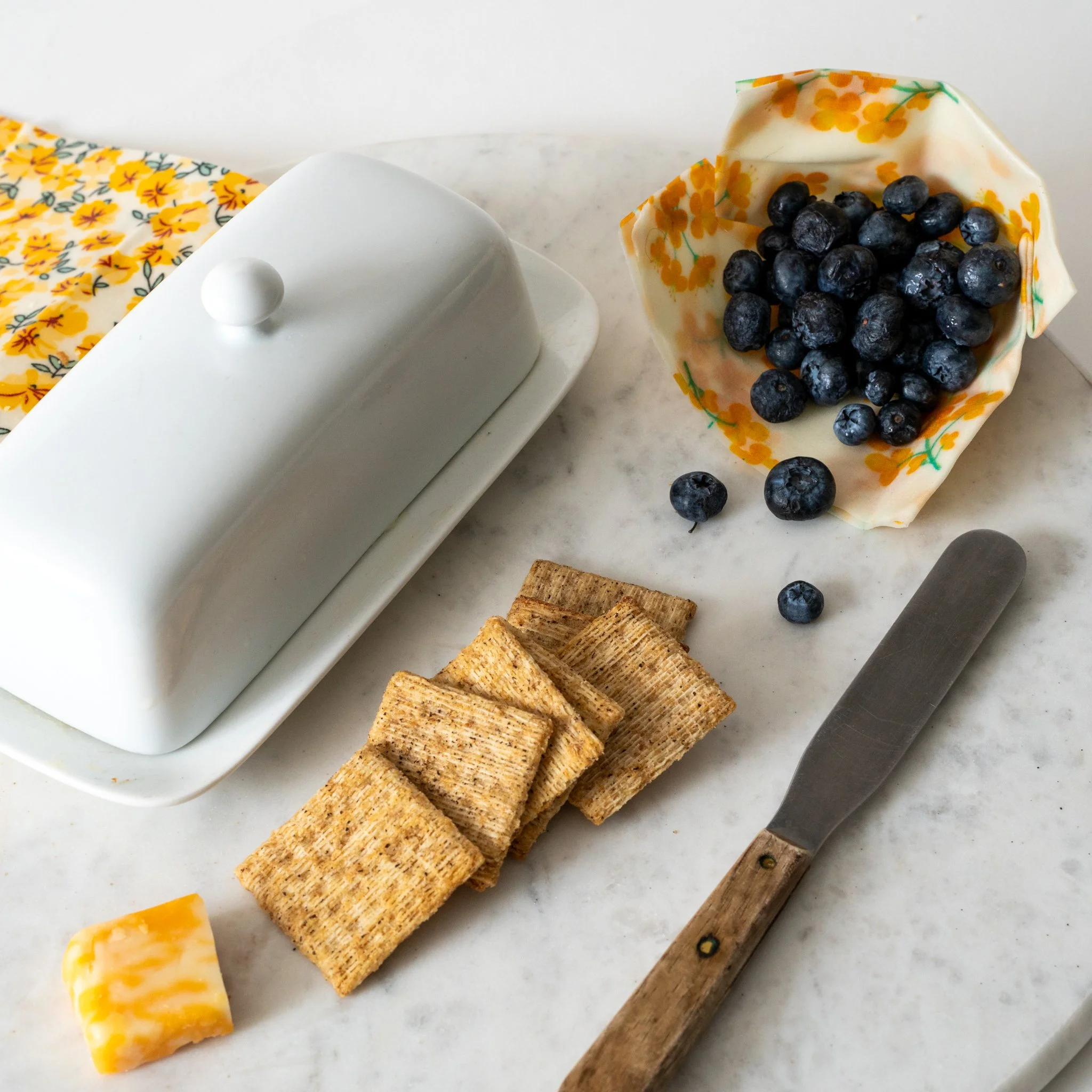 A marble countertop with a white butter dish, a small piece of yellow cheese, rectangular wheat crackers, a butter knife with a wooden handle, and a bowl filled with blueberries in a floral wrapper.