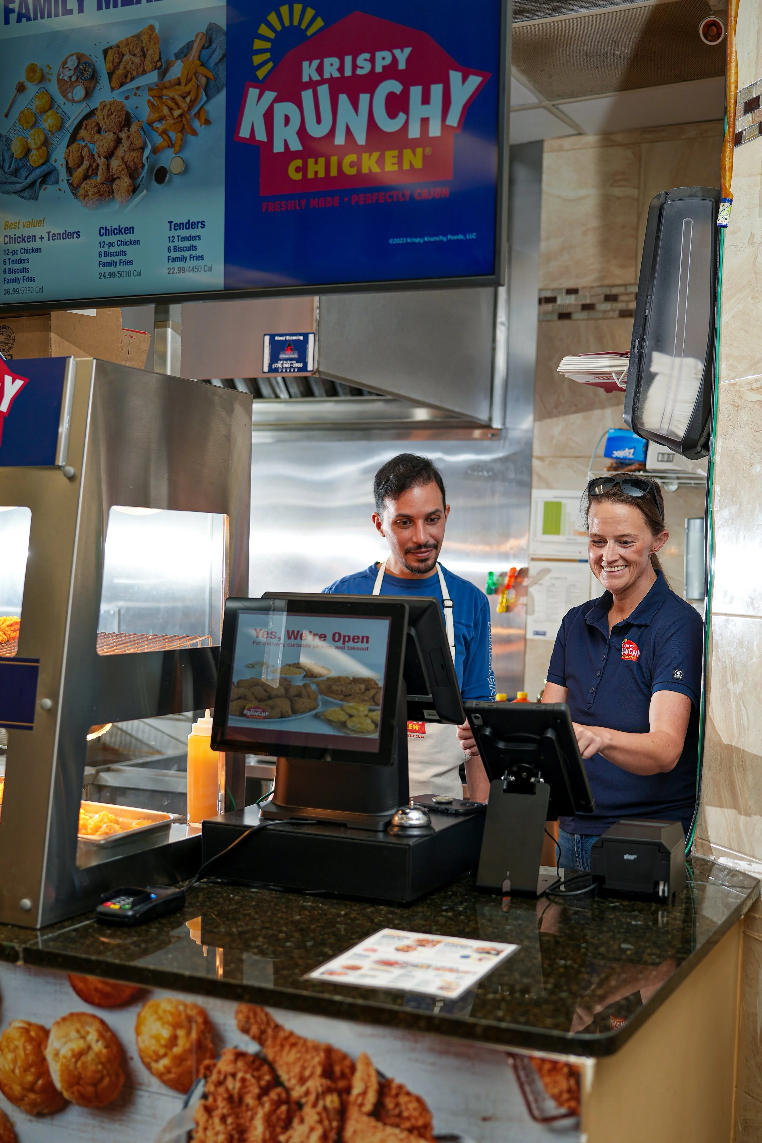 Two employees behind a counter at a fast food restaurant, with a large menu board above them displaying fried chicken options and a sign on the counter reading "Yes, We're Open."