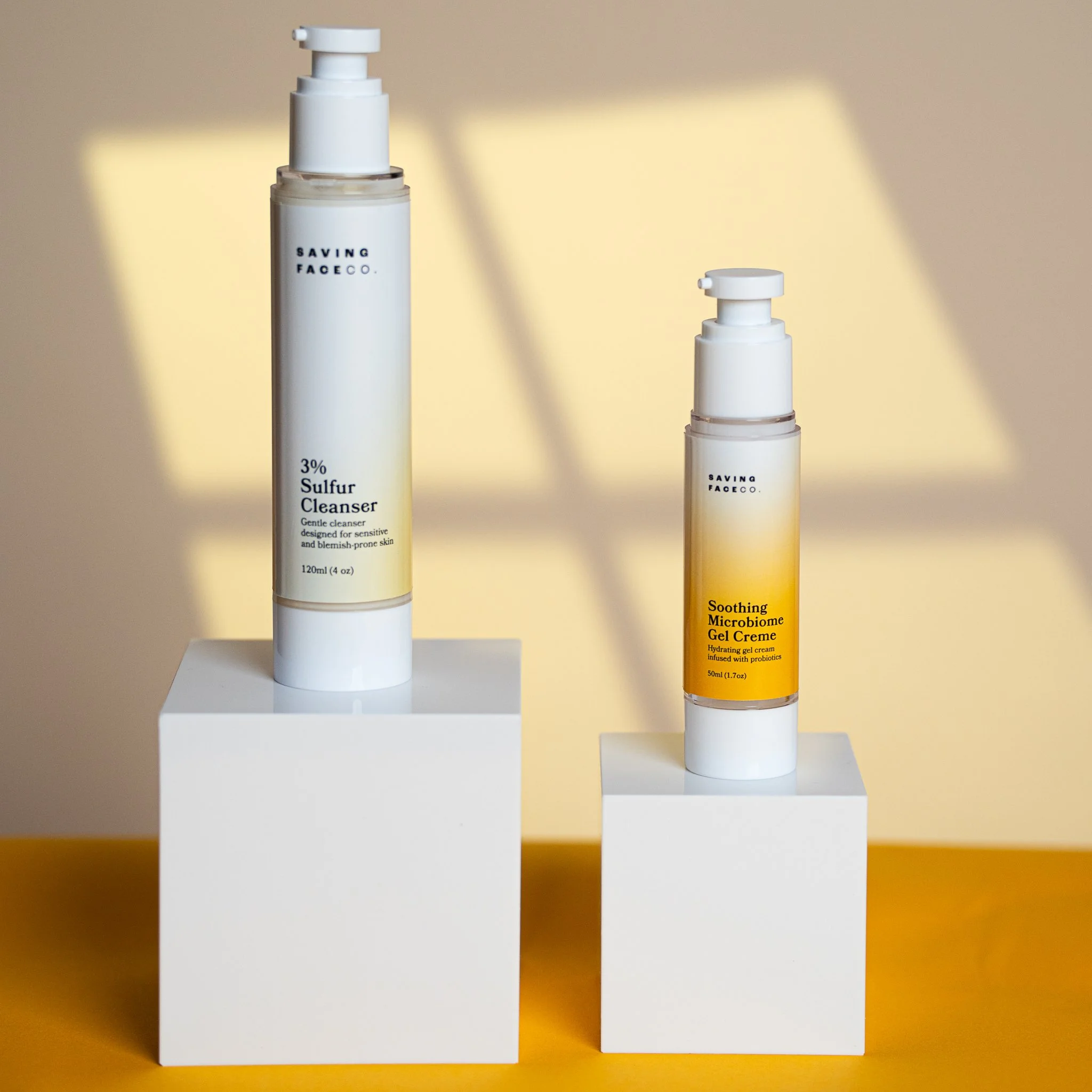 Two skincare bottles on white pedestals. The left bottle is taller with a label reading "Saving Face Co. 3% Sulfur Cleanser." The right bottle, shorter, labeled "Saving Face Co. Soothing Microbiome Gel Creme." The background has a warm light with shadows.