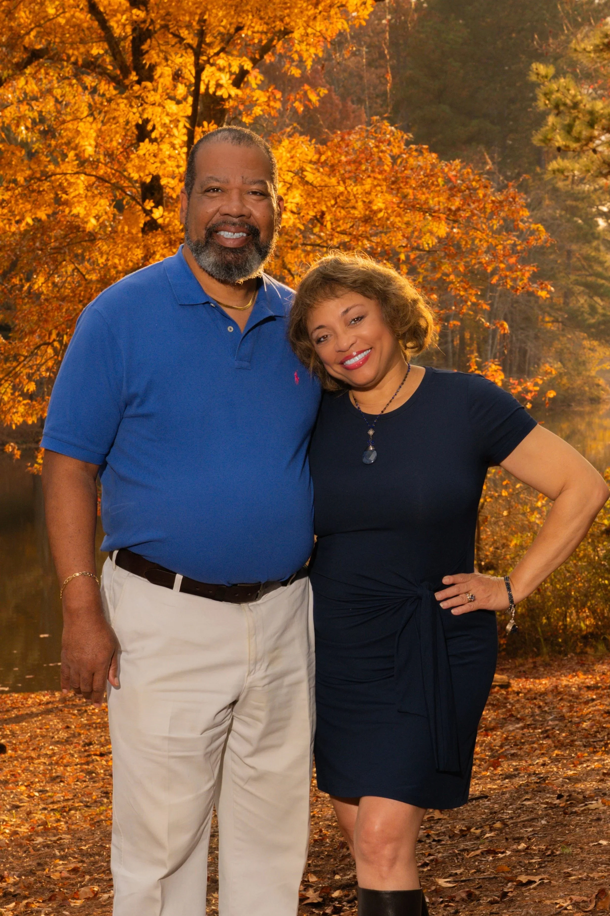 A smiling man and woman standing outdoors in front of autumn trees with orange and yellow leaves, near a body of water.