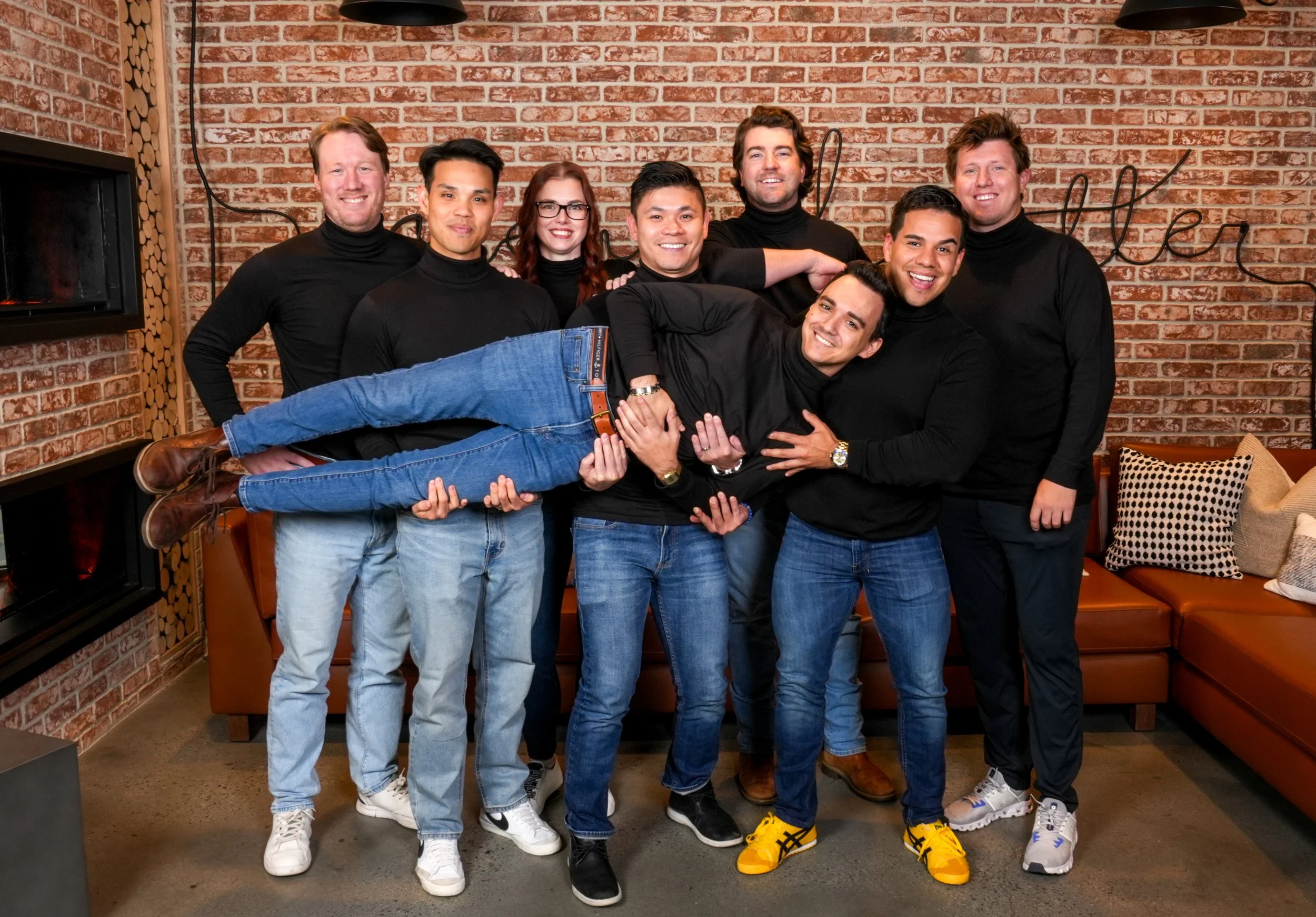 A group of eight people, all wearing black turtleneck sweaters, is posing together in a living room with a brick wall background. One person is being held horizontally across the others' laps and arms. They are smiling and appear to be having fun.