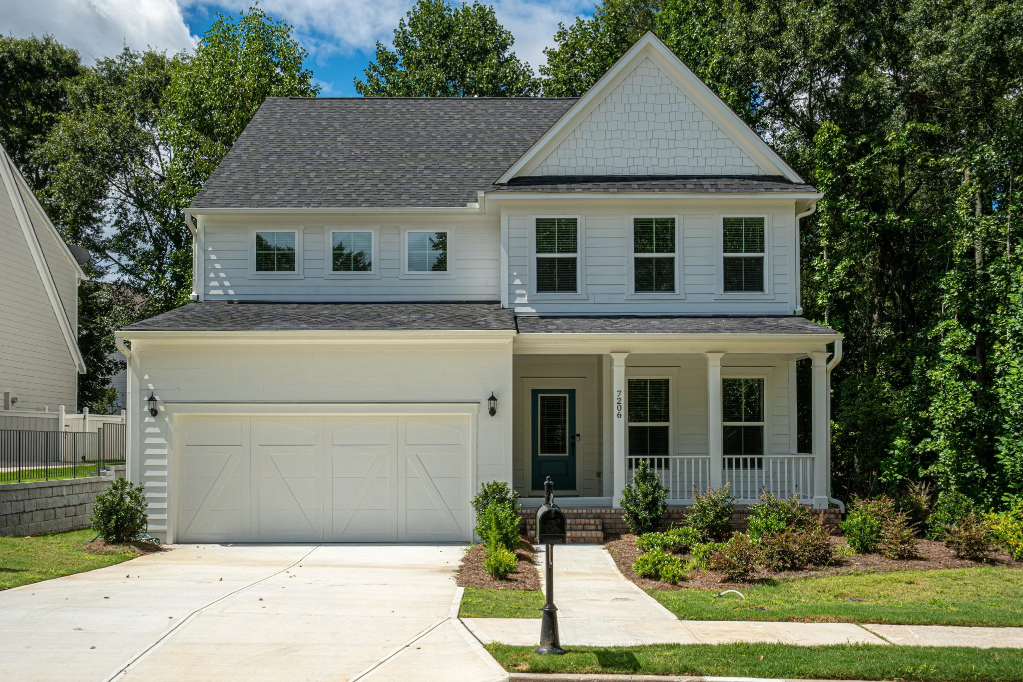 A two-story white house with a dark gray roof, front porch with white railing, and a green door. Well-maintained lawn and bushes in front, driveway on the left, mailbox near sidewalk, surrounded by green trees under a partly cloudy sky.