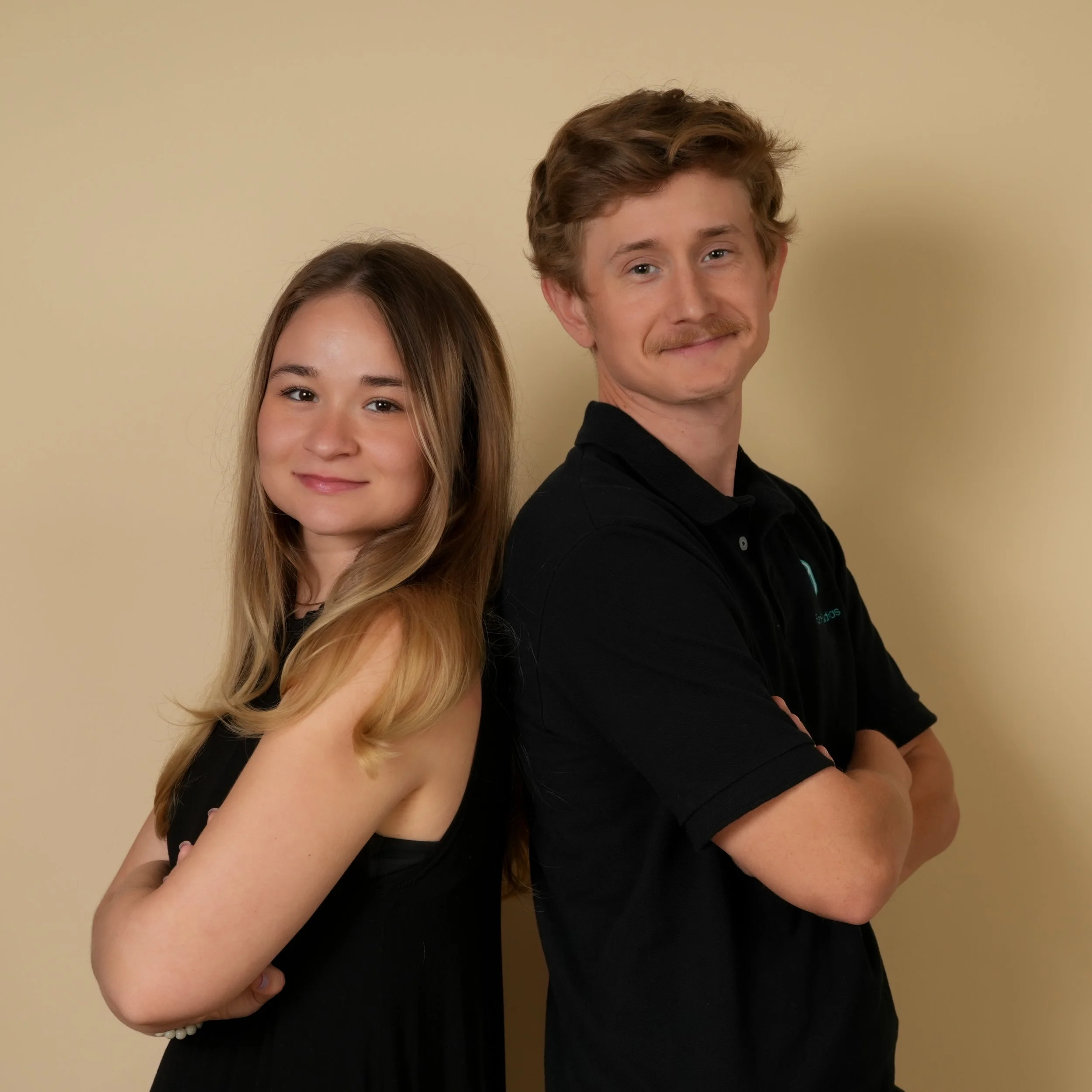 A young woman and a young man stand back to back with their arms crossed against a plain beige wall, smiling at the camera.