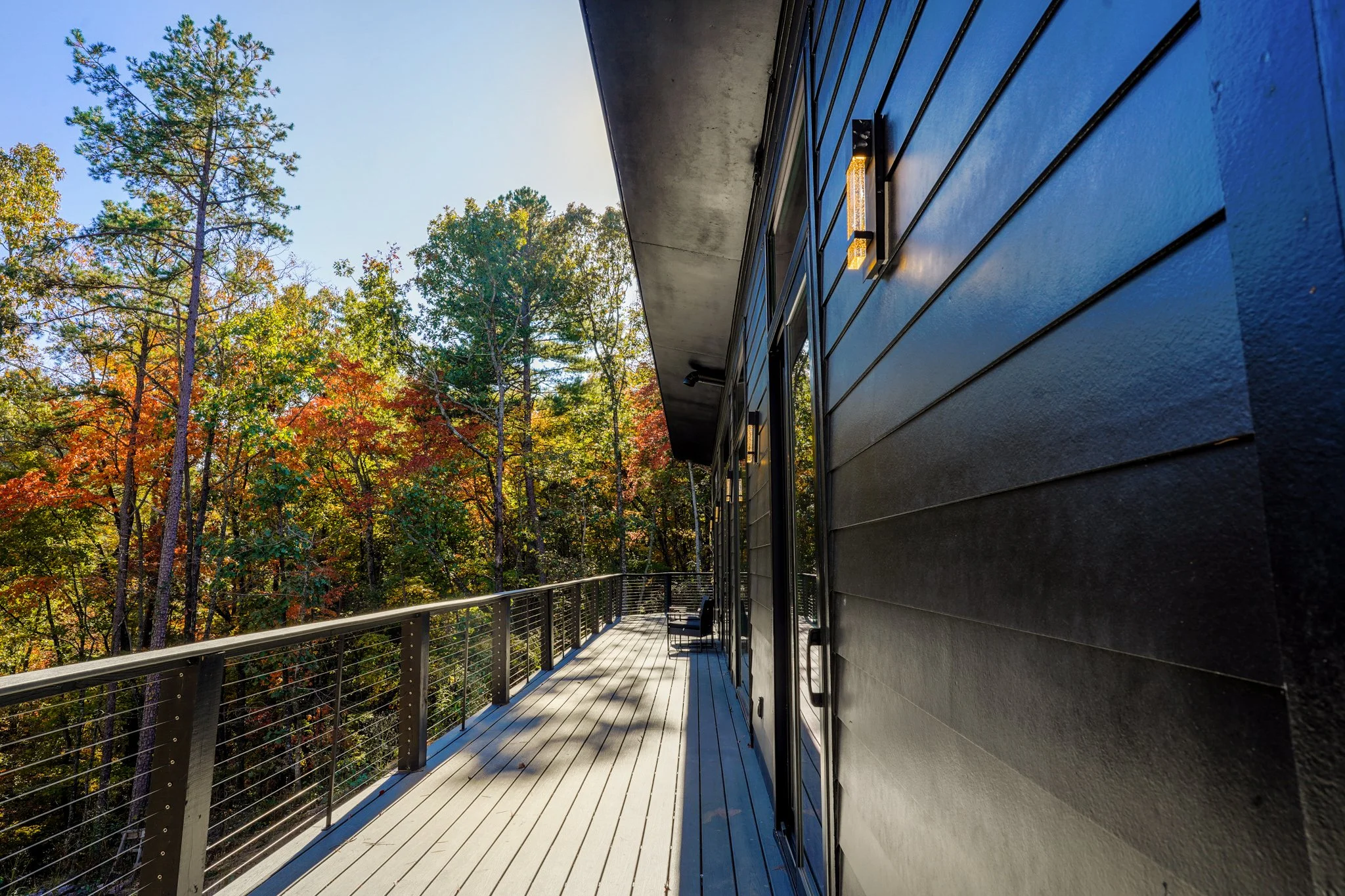 A modern balcony with a wooden floor and metal railing attached to a black house, overlooking a forest with trees showing autumn colors, under a clear sky.