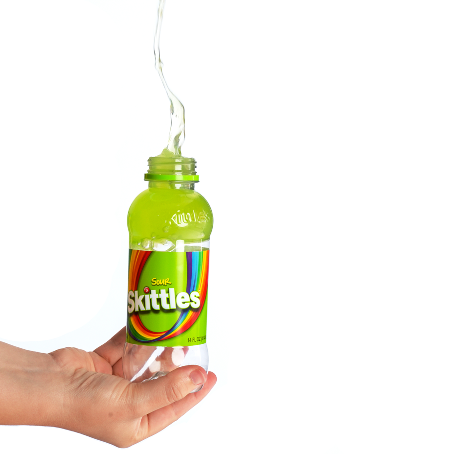 A hand holding a clear plastic bottle with a green label and label reads 'Skittles Sour' with colorful rainbow design, and water is being poured into it.