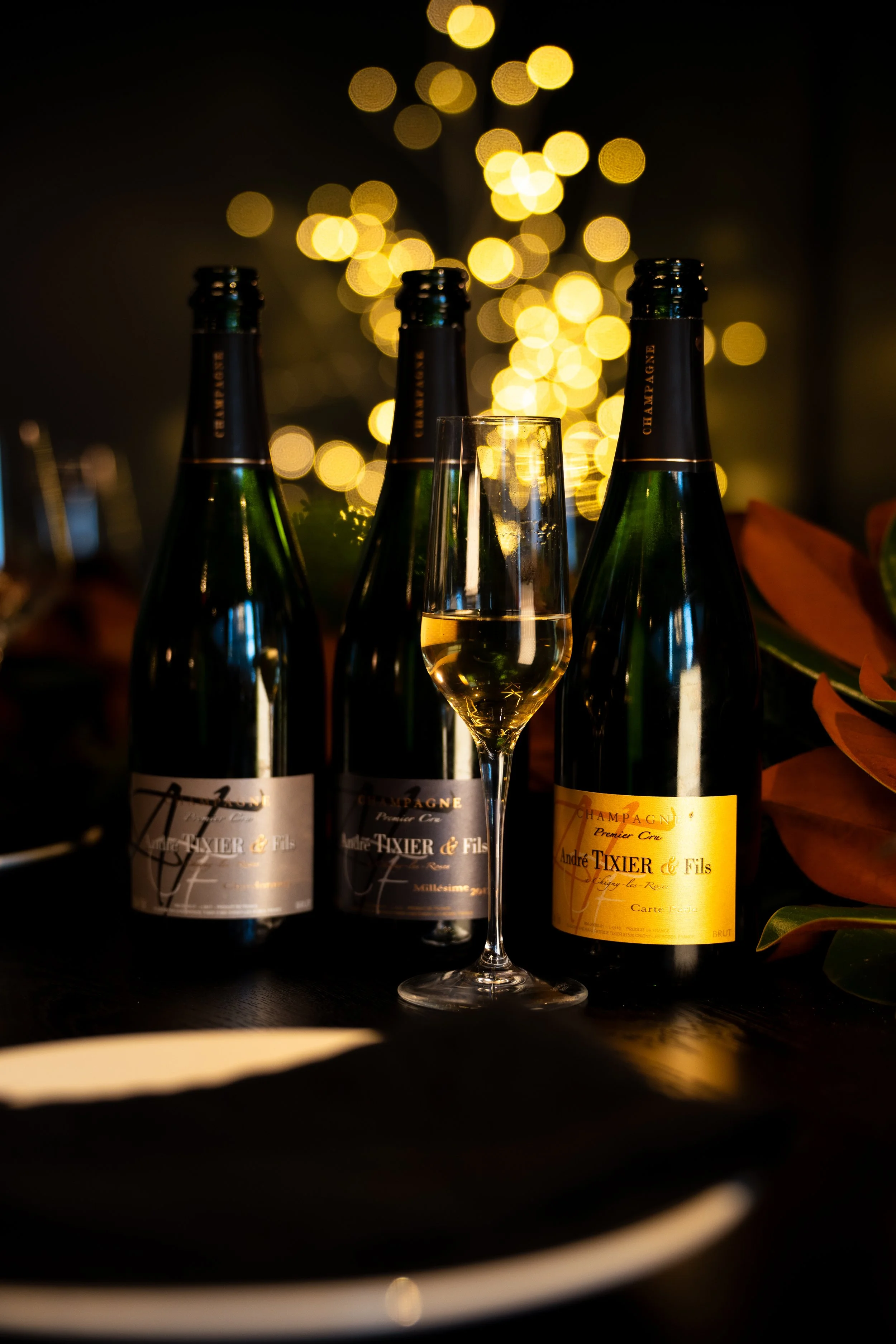 Three bottles of champagne and a champagne flute filled with champagne on a dark surface with blurred warm yellow bokeh lights in the background, surrounded by green and orange foliage.