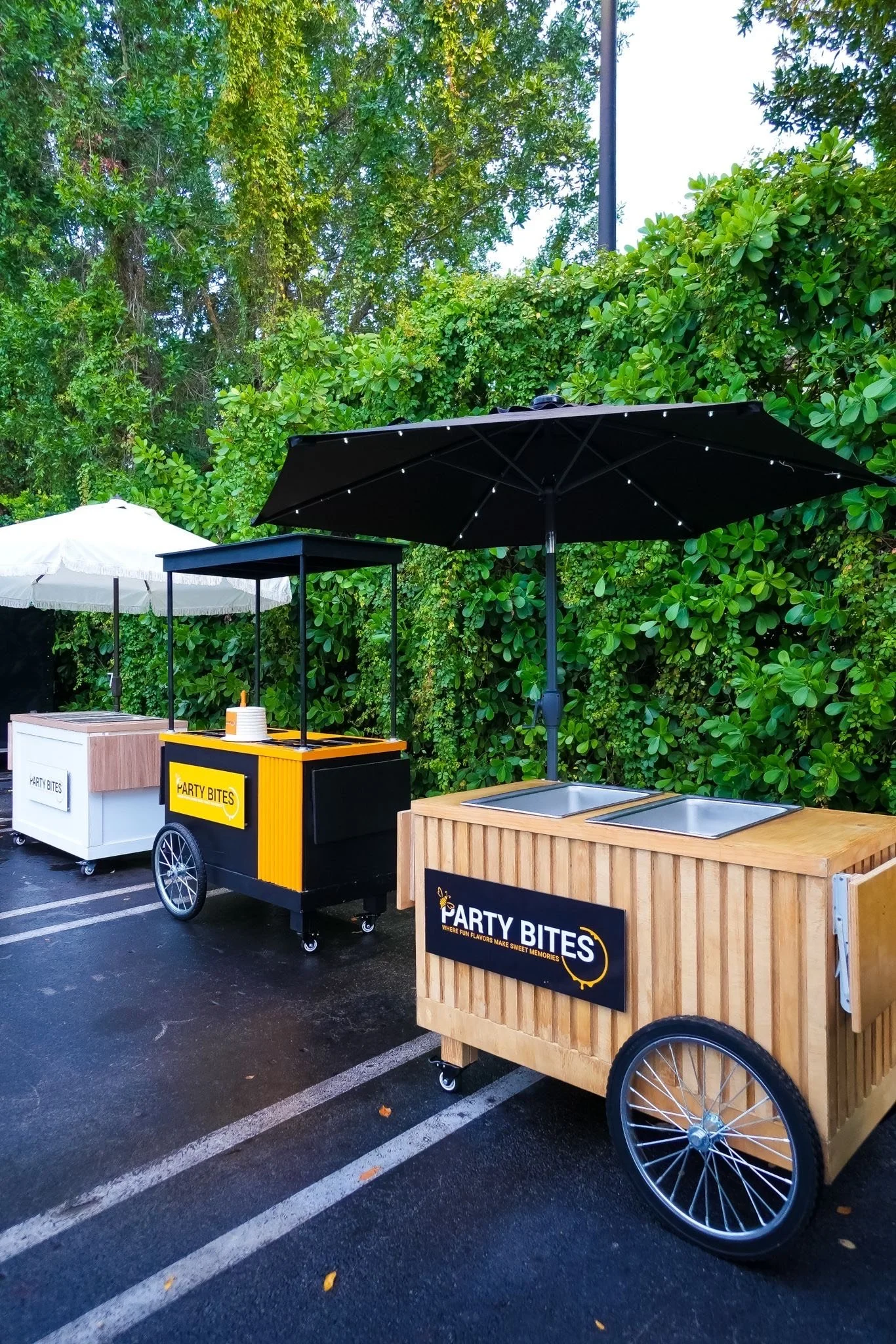 Three beverage carts with umbrellas in a parking lot, one black and yellow branded 'Party Bites' and two white carts, surrounded by lush green trees.