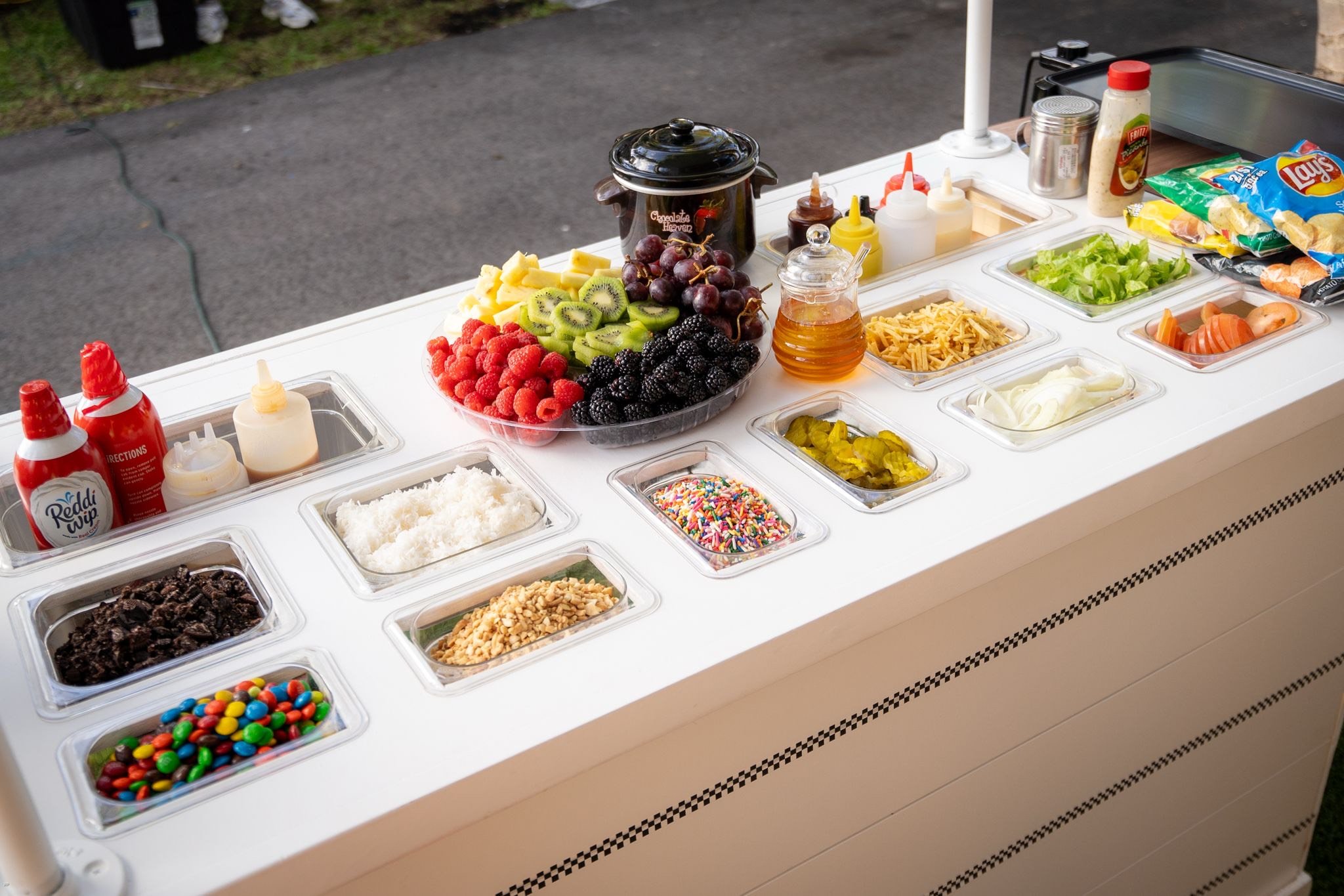 Ice cream sundae bar with various toppings including candies, fruits, sprinkles, nuts, and whipped cream, set up on a white cart outdoors.