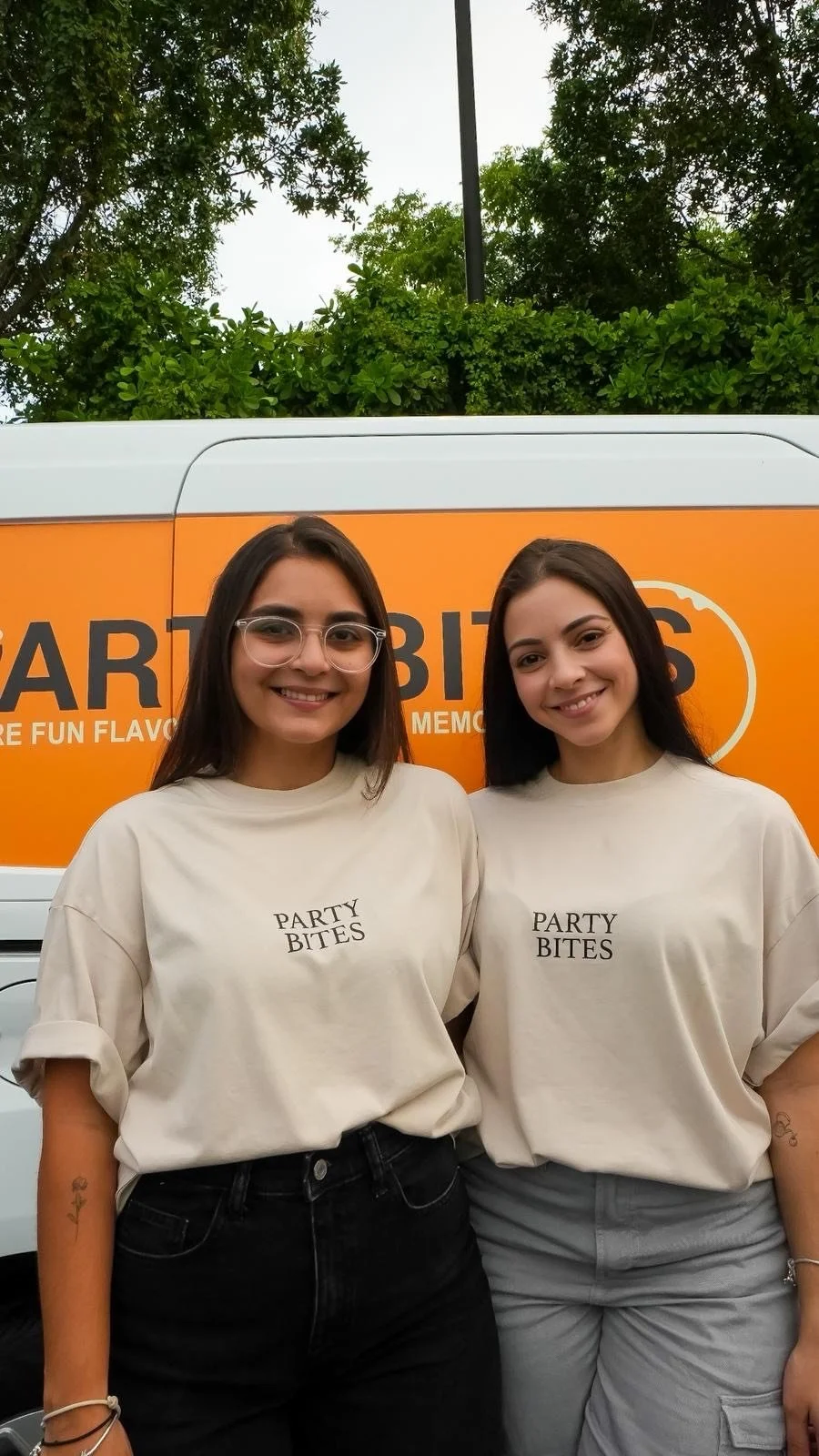 Two women smiling both team for Party Bites Mia, wearing matching beige T-shirts with 'PARTY BITES' printed on them, standing in front of an orange mobile food stand with the words 'PARTY BITES' on it and some greenery above.