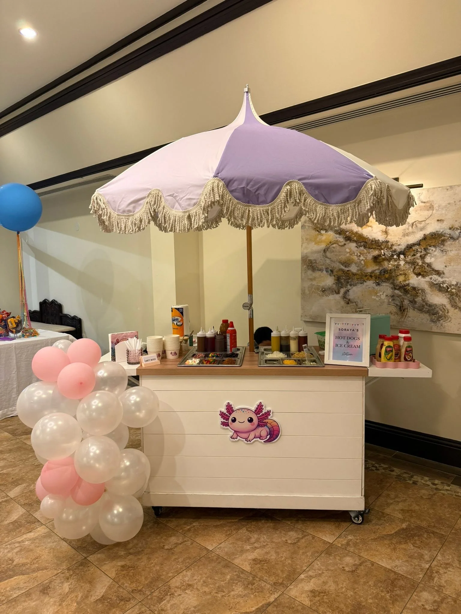 A small outdoor-style stand with a large purple and white umbrella. The stand has condiments, cups. It's a Party Bites MIA event.  There are pink and white balloons attached to the stand, and an abstract painting on the wall behind it.