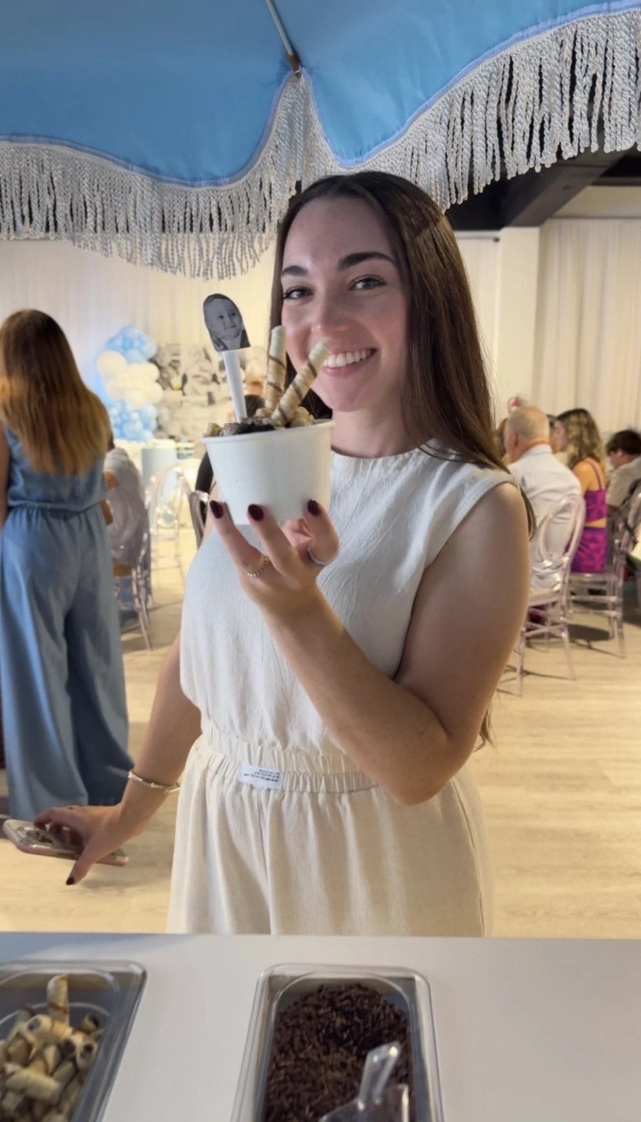 A woman smiling and holding a cup of ice cream topped with cookies and wafer sticks at a party or celebration, with other guests in the background.