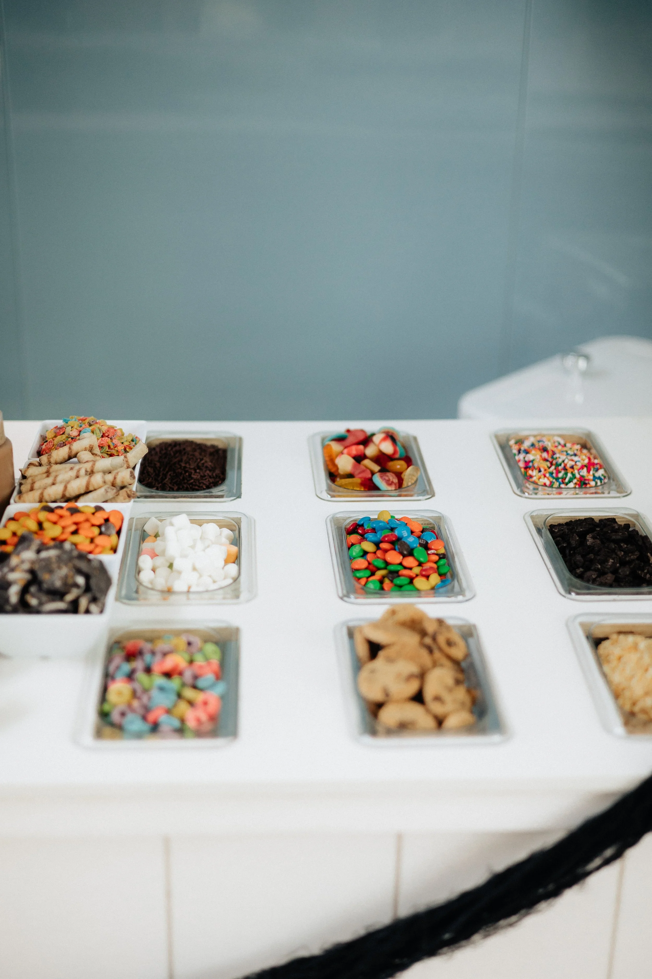A variety of colorful candies and sprinkles in metal trays on a white countertop.