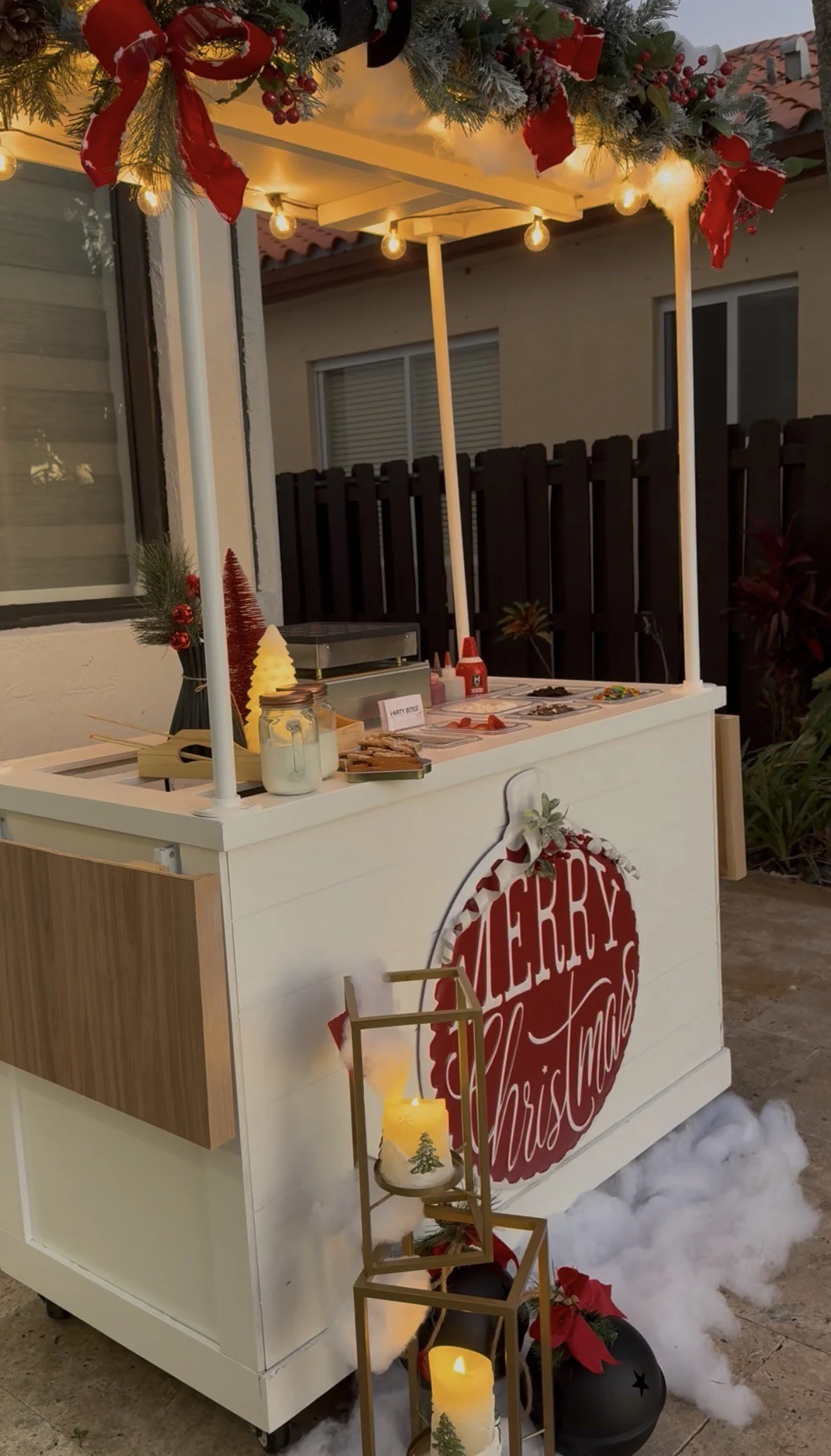 Christmas-themed outdoor vendor cart decorated with holiday ornaments, garlands, and lights, with candles, poinsettias, and a sign that says "Merry Christmas".