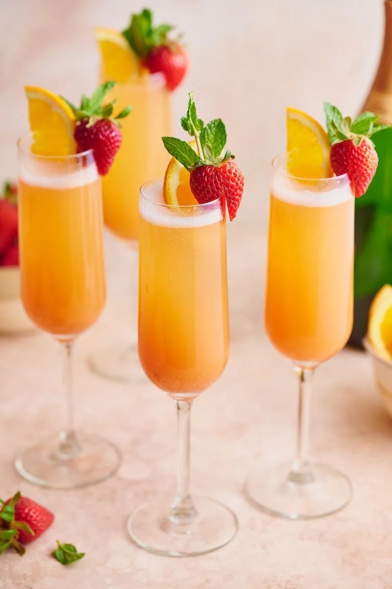 Three glasses of peach-colored champagne cocktails garnished with strawberries, orange slices, and mint leaves.