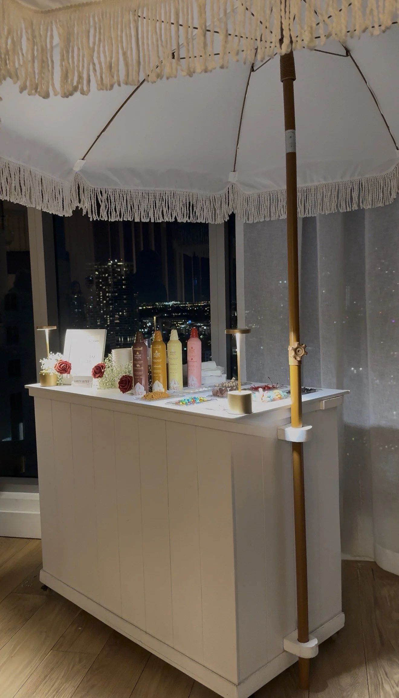 Nighttime view of a decorated party table with colorful bottles, flowers, and treats, covered by a large white umbrella with tasseled edges, near a window showing city lights.