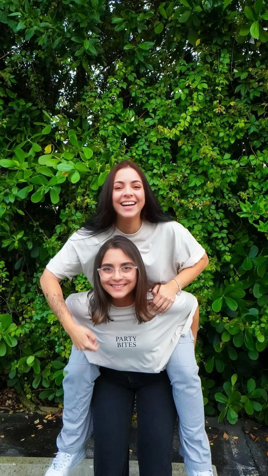Two young women smiling, one giving the other a piggyback ride in front of lush green shrubbery.