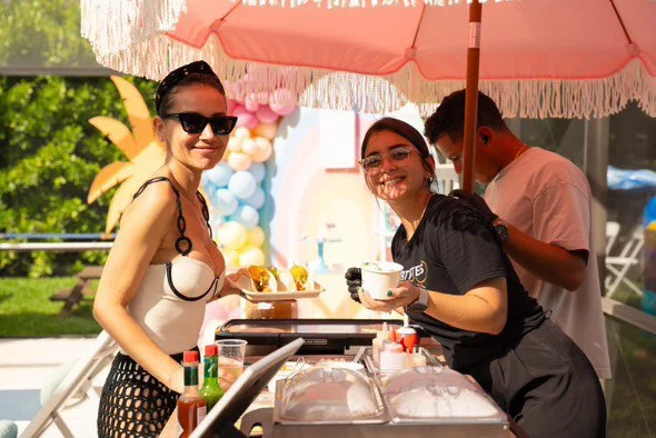 Three people working at a food stall outdoors under a pink umbrella, with balloons and greenery in the background. Two women are smiling; one is wearing sunglasses and holding a plate, the other is holding a cup. A man is in the background.