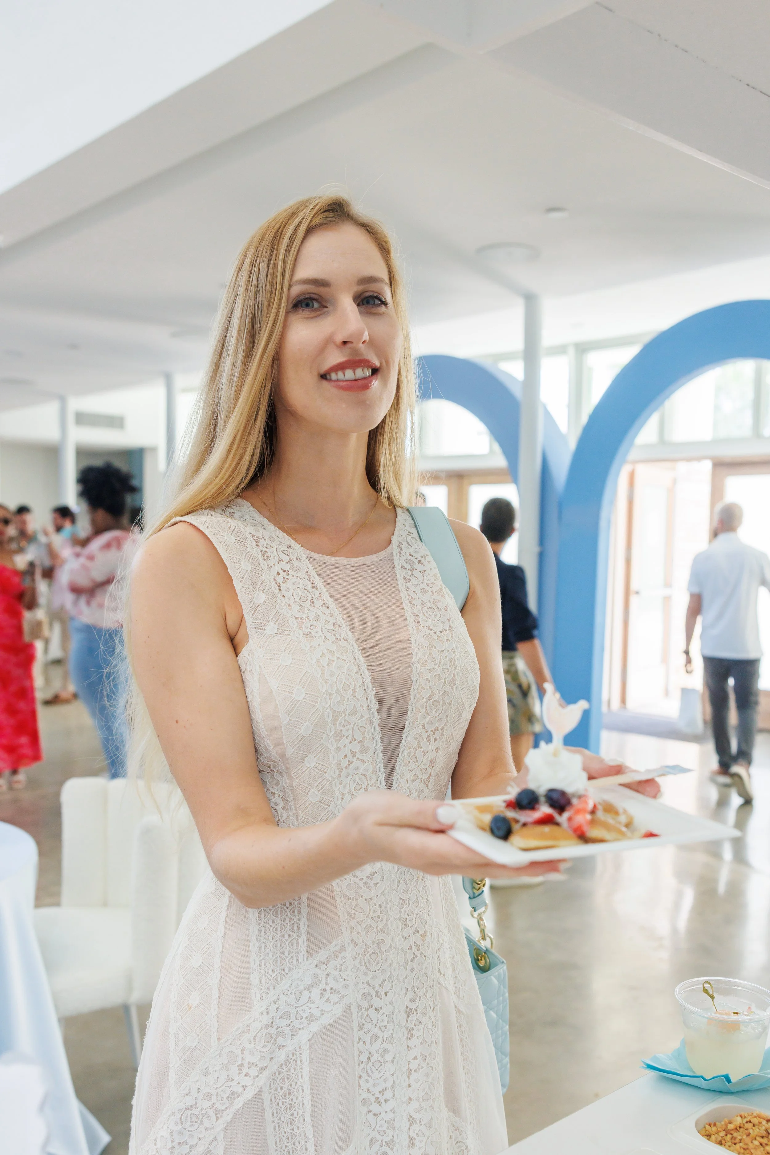 A woman with long blonde hair in a sleeveless beige lace dress holding a plate of dessert, inside a bright, modern venue with blue decorative arches and other people in the background.