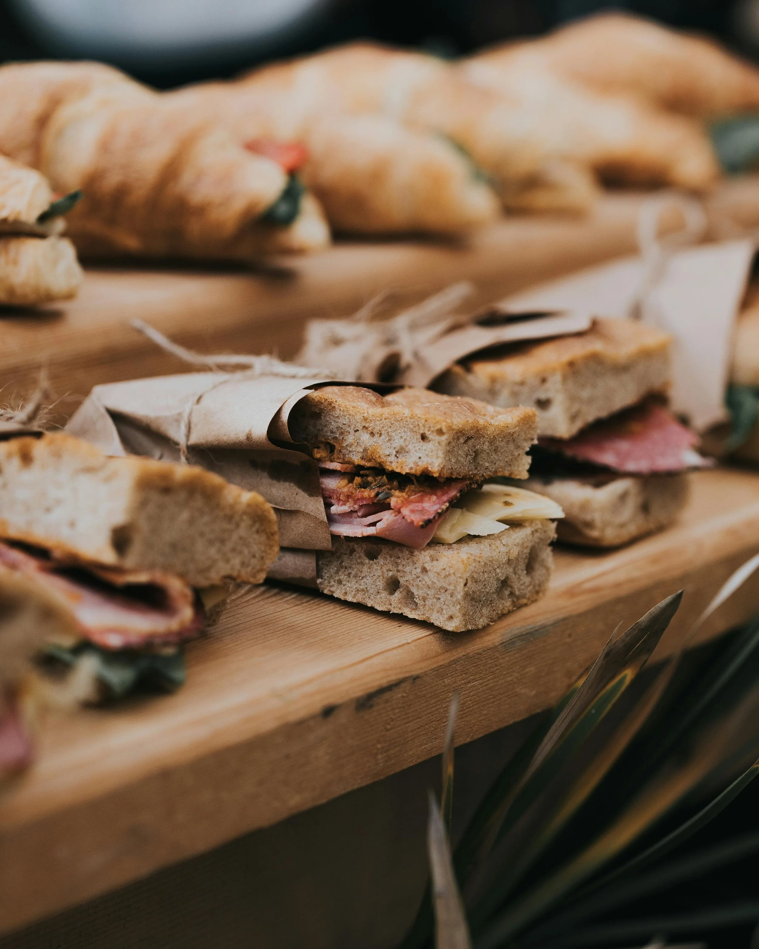 Close-up of sandwiches wrapped in paper and tied with string, displayed on a wooden serving board, with croissants in the background.