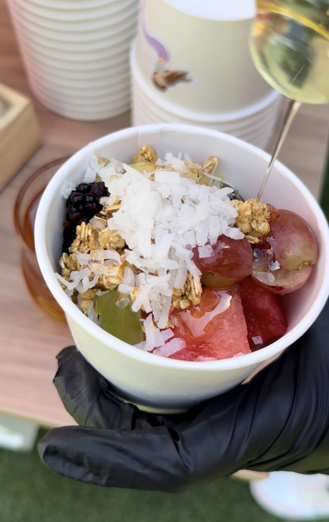A cup of yogurt topped with granola, coconut flakes, grapes, and blackberries, with a glass of white wine in the background, held by a person wearing black gloves.