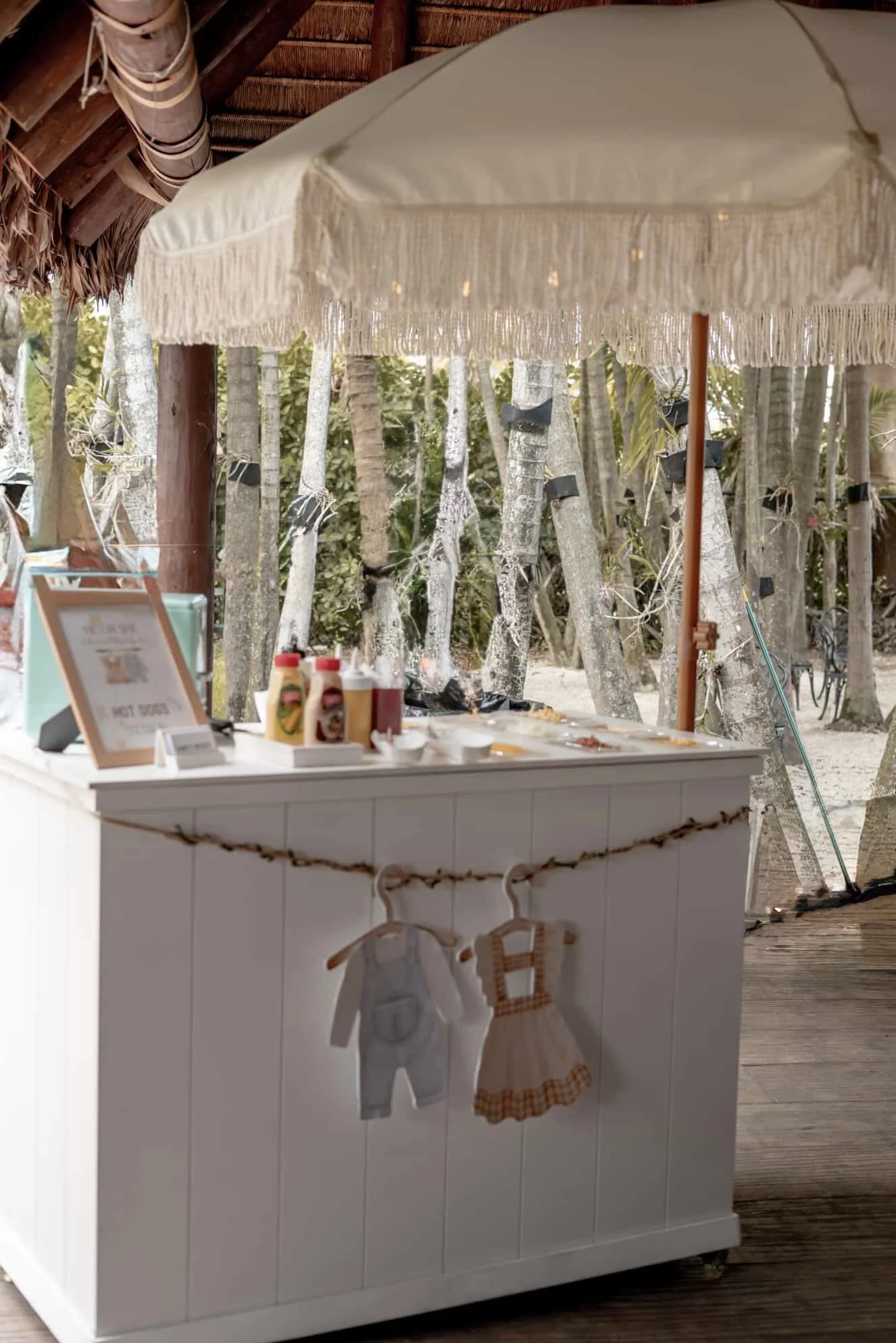 Beachside food stand with a white counter, condiments, and a framed sign, under a thatched roof with flowy fringe, near trees with black tape and beach surroundings.