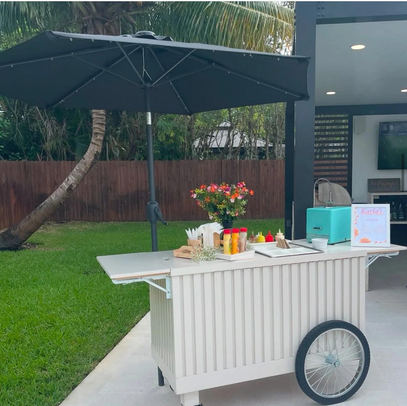 A white mobile food or drink cart with a black umbrella, set up outdoors on a patio. The cart has condiments, napkins, flowers, and a small blue appliance, with a table sign reading 'Karla's'. There is a grassy area, a wooden fence, and tropical plan