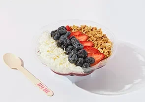 Bowl of yogurt topped with blueberries, strawberries, granola, and shredded coconut, with a wooden spoon nearby