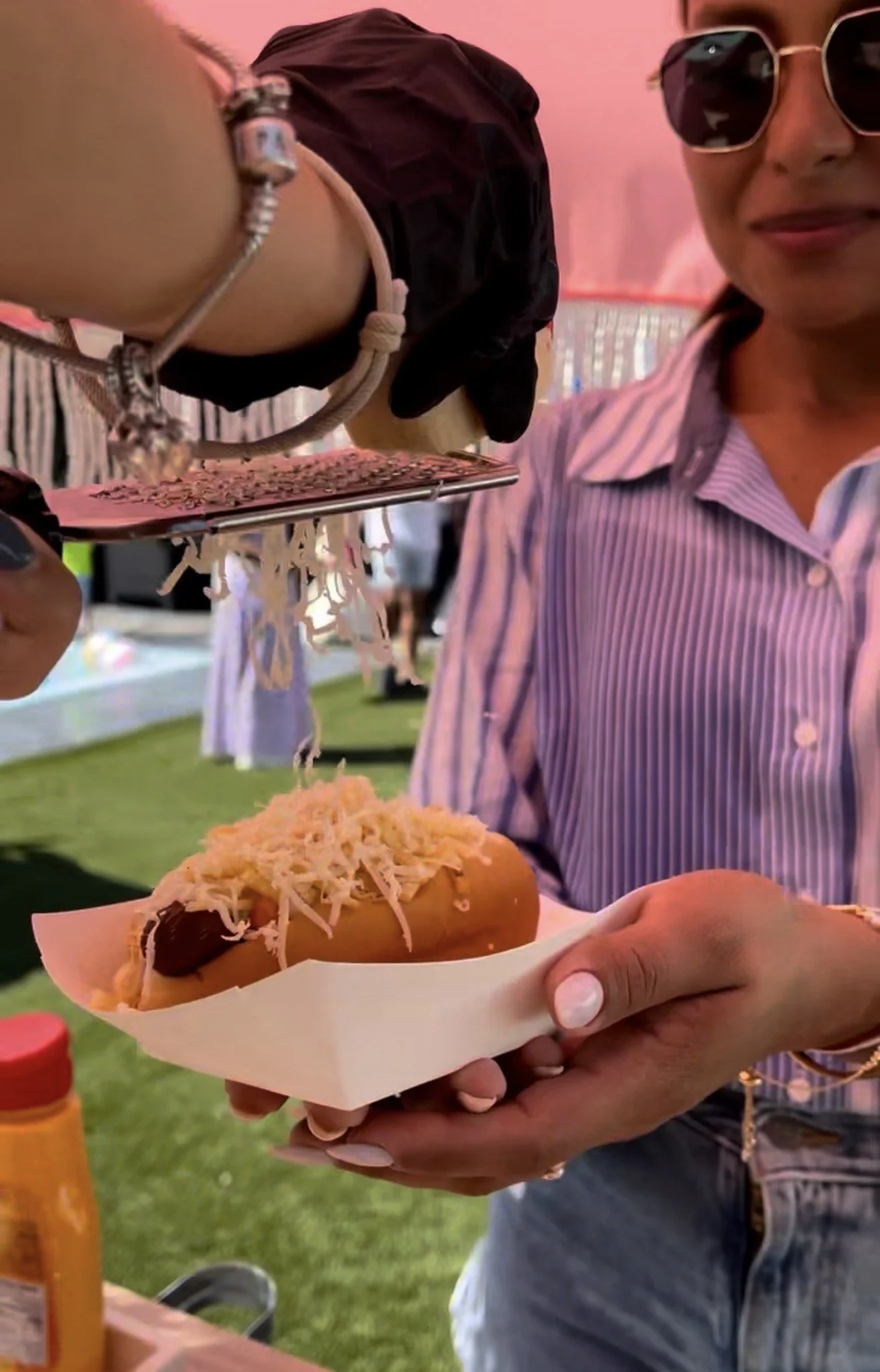 A woman wearing sunglasses and a pink striped shirt holding a hot dog topped with shredded cheese and other toppings at an outdoor event.