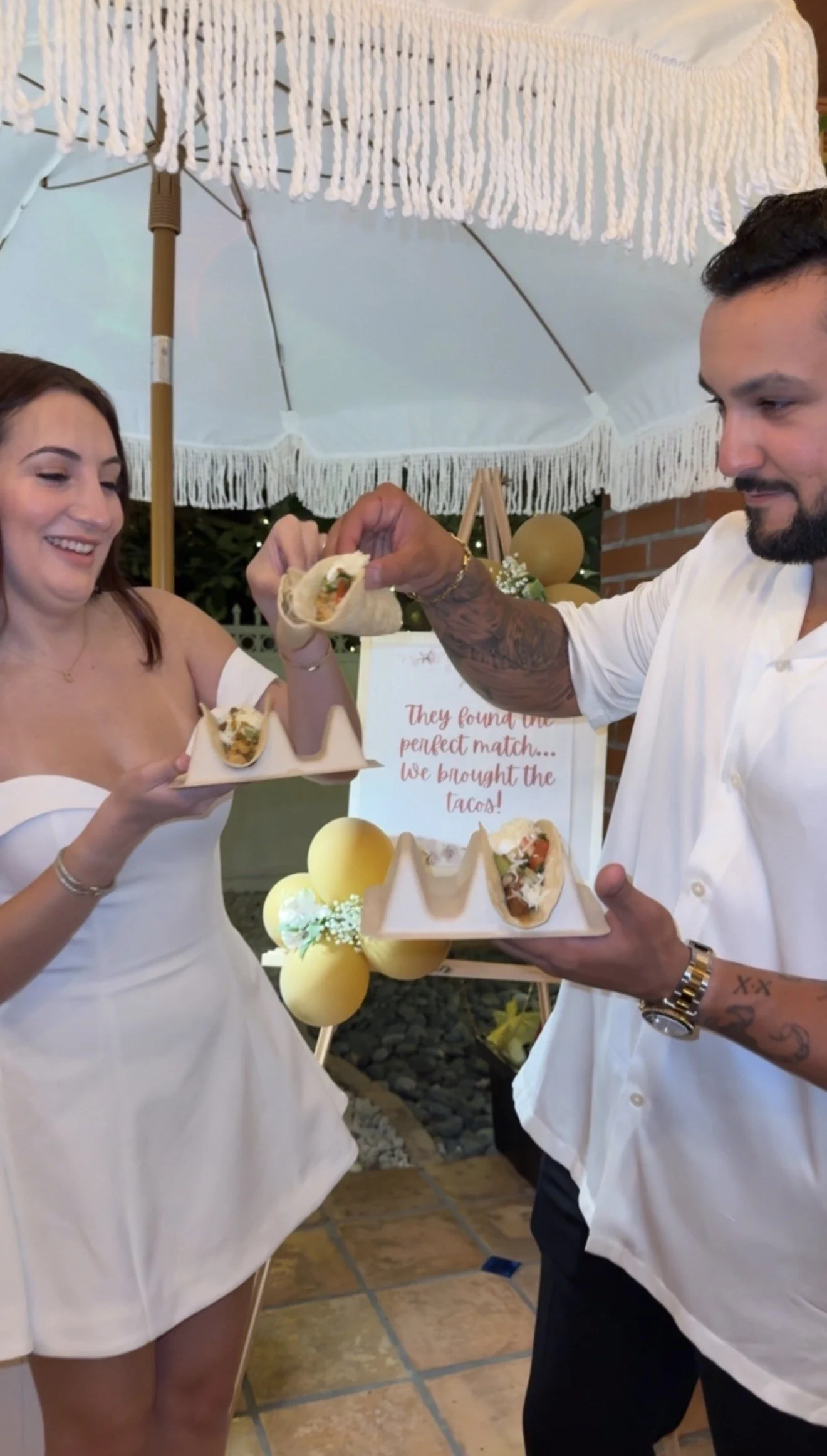 A couple at a celebration sharing tacos under a white parasol, with a sign in the background reading 'They found their perfect match... We brought the tacos!', decorated with yellow balloons.