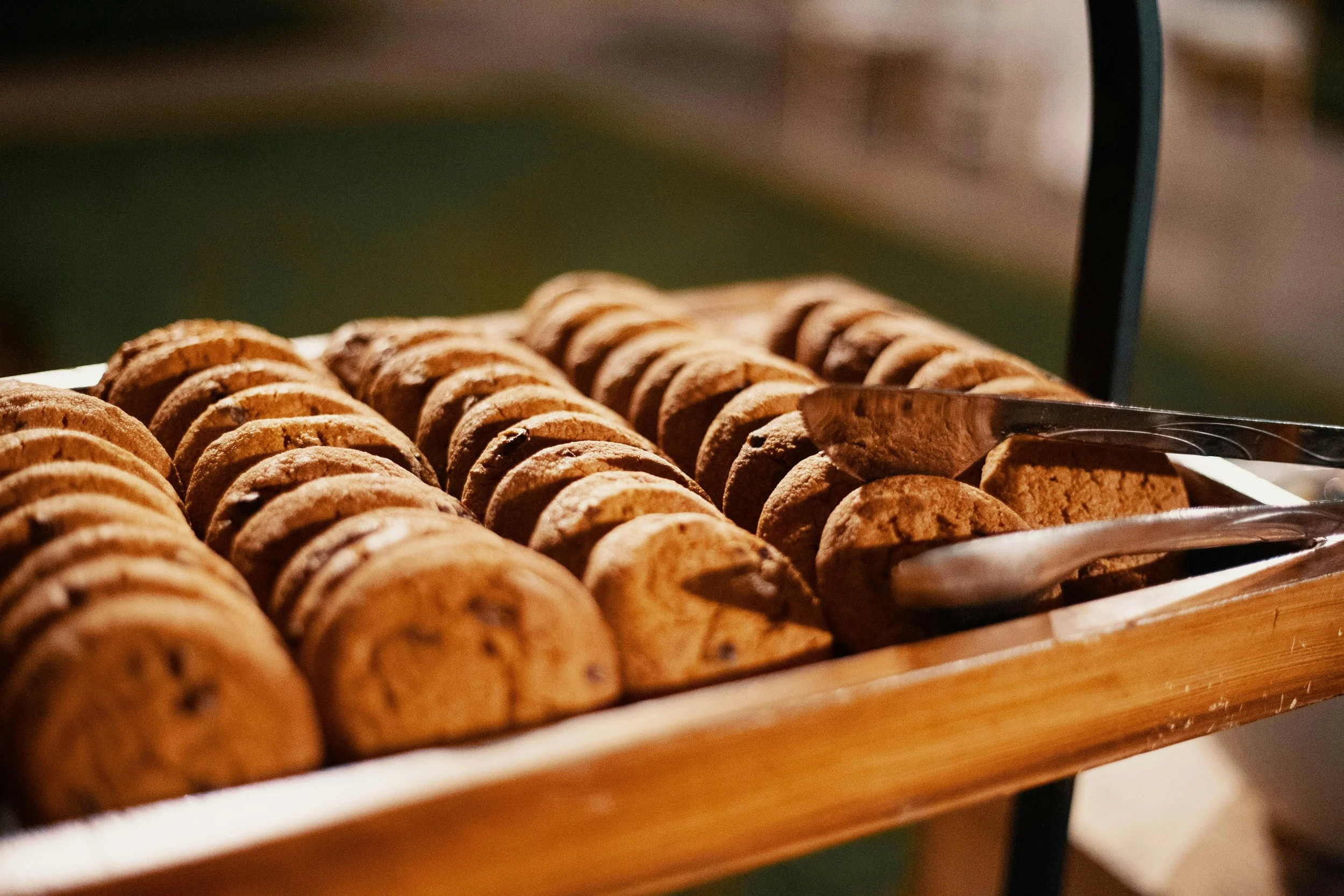 A tray of chocolate chip cookies arranged in rows, with tongs placed on the cookies for serving.